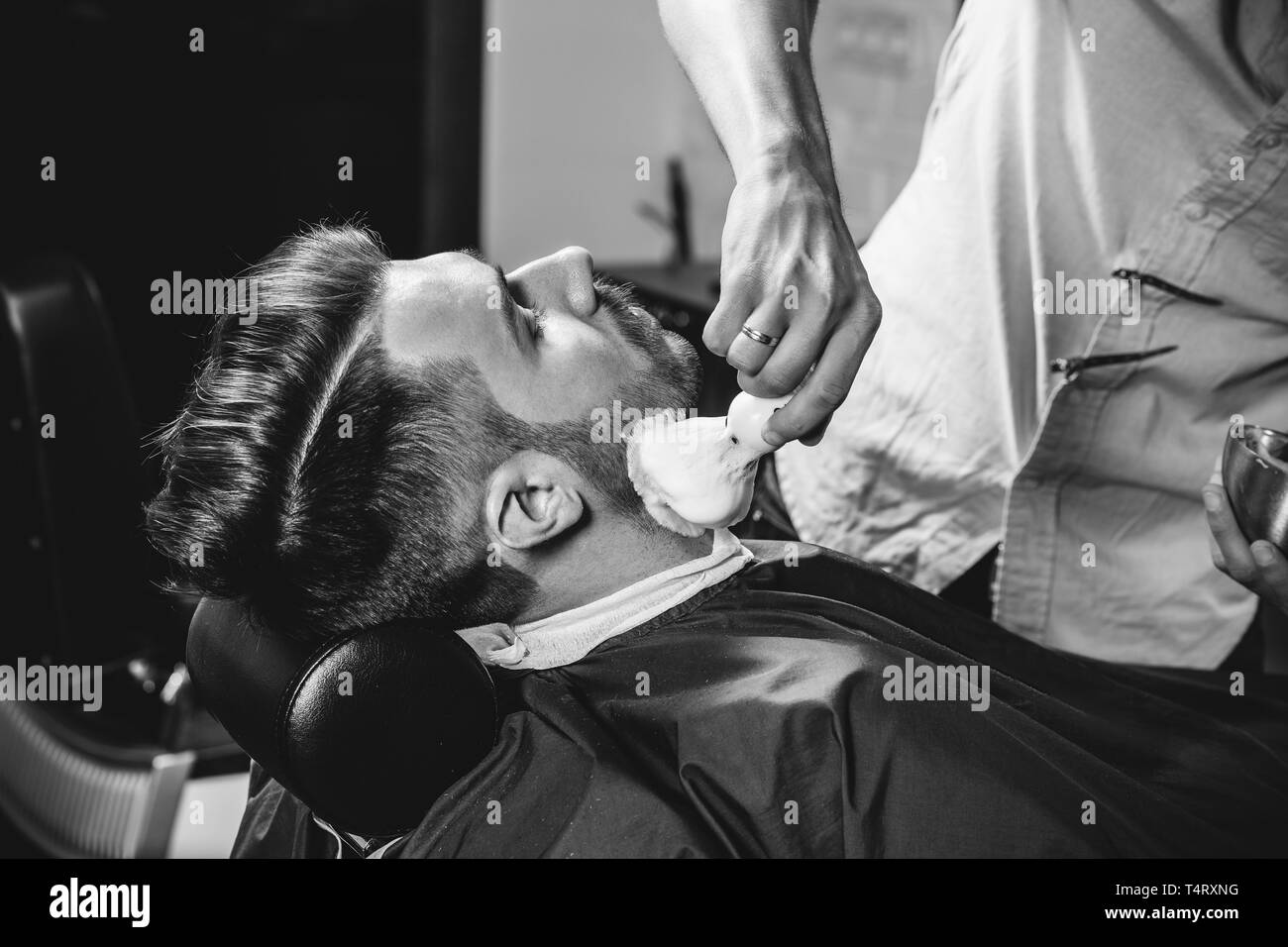 Client durante la rasatura della barba in barberia. Giovani bel barbiere e attraente uomo barbuto. Nero e bianco o incolore foto. Acconciatura, salone di bellezza e parrucchiere, barbiere, il concetto di stile di vita. Caucasian modelli maschili. Foto Stock