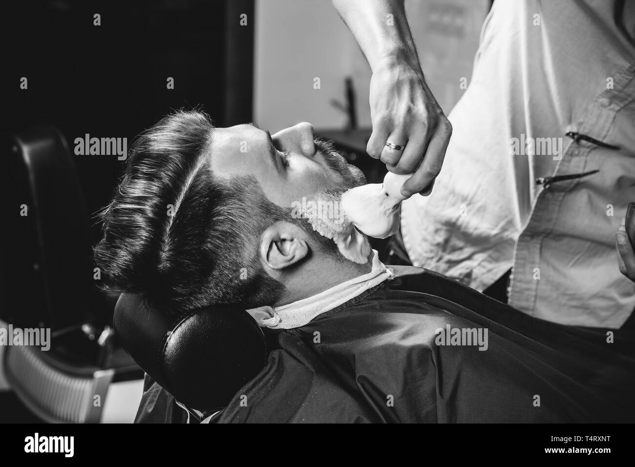 Client durante la rasatura della barba in barberia. Giovani bel barbiere e attraente uomo barbuto. Nero e bianco o incolore foto. Acconciatura, salone di bellezza e parrucchiere, barbiere, il concetto di stile di vita. Caucasian modelli maschili. Foto Stock
