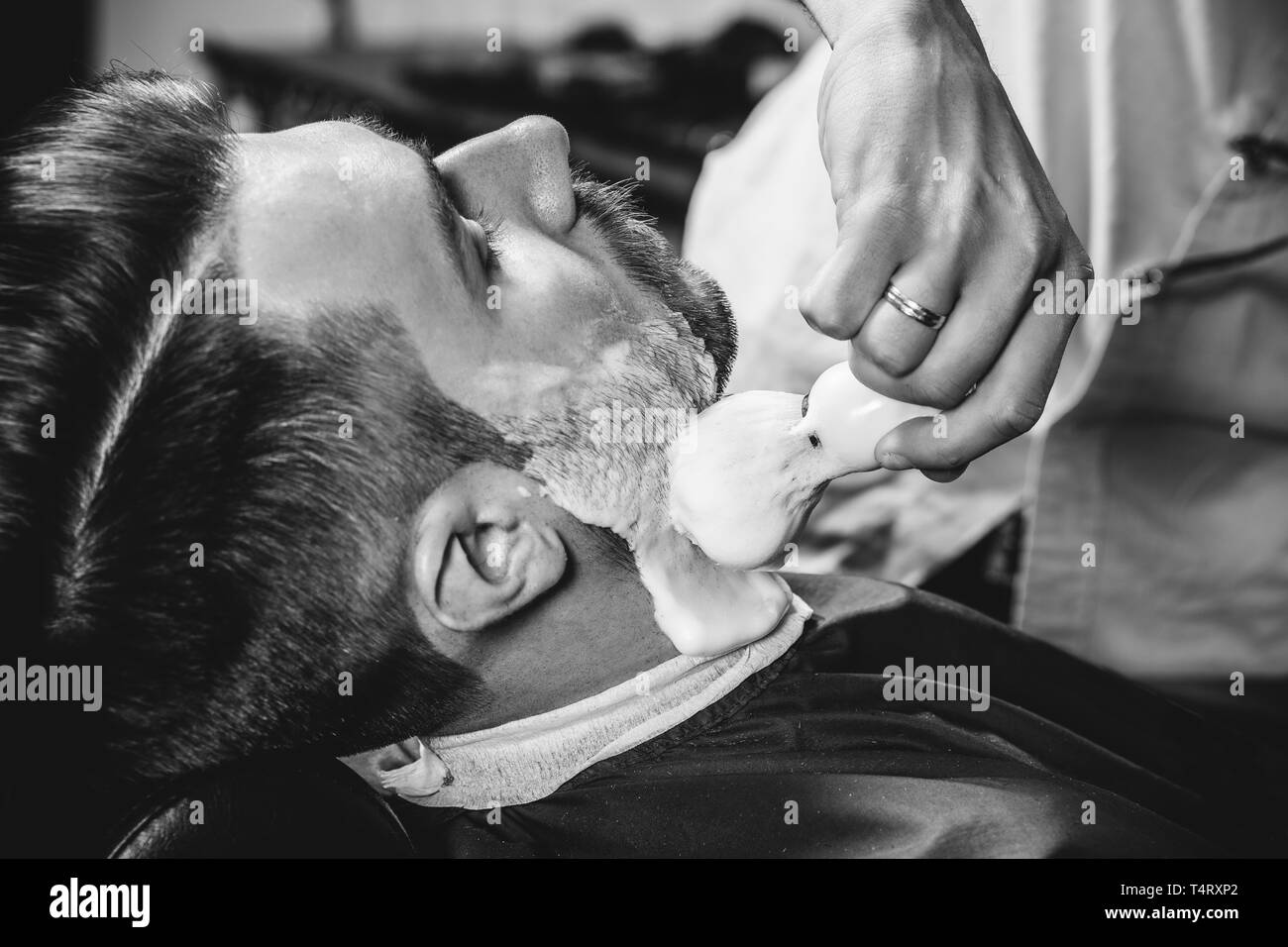 Client durante la rasatura della barba in barberia. Giovani bel barbiere e attraente uomo barbuto. Nero e bianco o incolore foto. Acconciatura, salone di bellezza e parrucchiere, barbiere, il concetto di stile di vita. Caucasian modelli maschili. Foto Stock