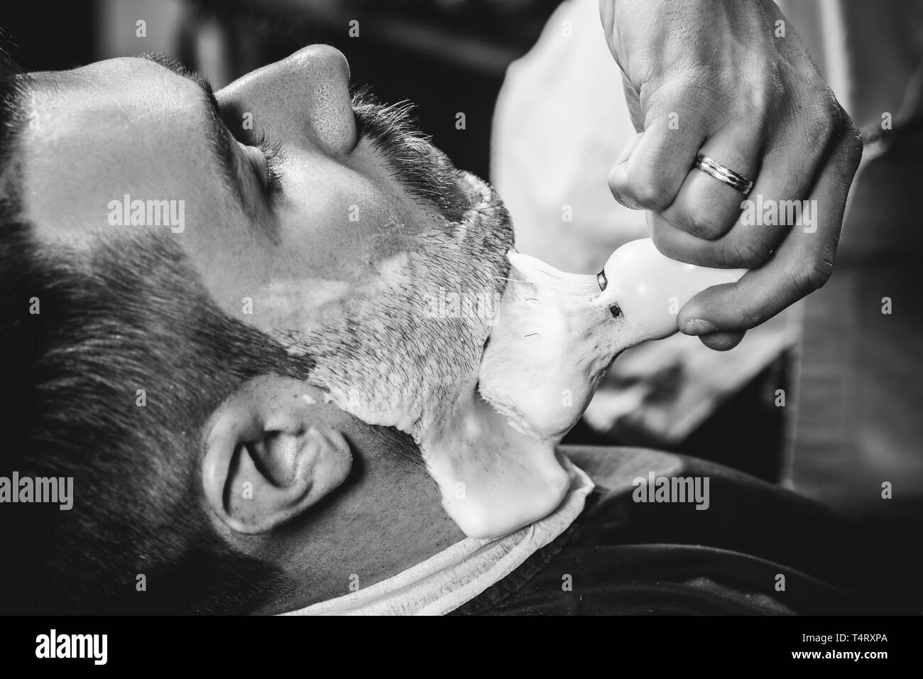 Client durante la rasatura della barba in barberia. Giovani bel barbiere e attraente uomo barbuto. Nero e bianco o incolore foto. Acconciatura, salone di bellezza e parrucchiere, barbiere, il concetto di stile di vita. Caucasian modelli maschili. Foto Stock