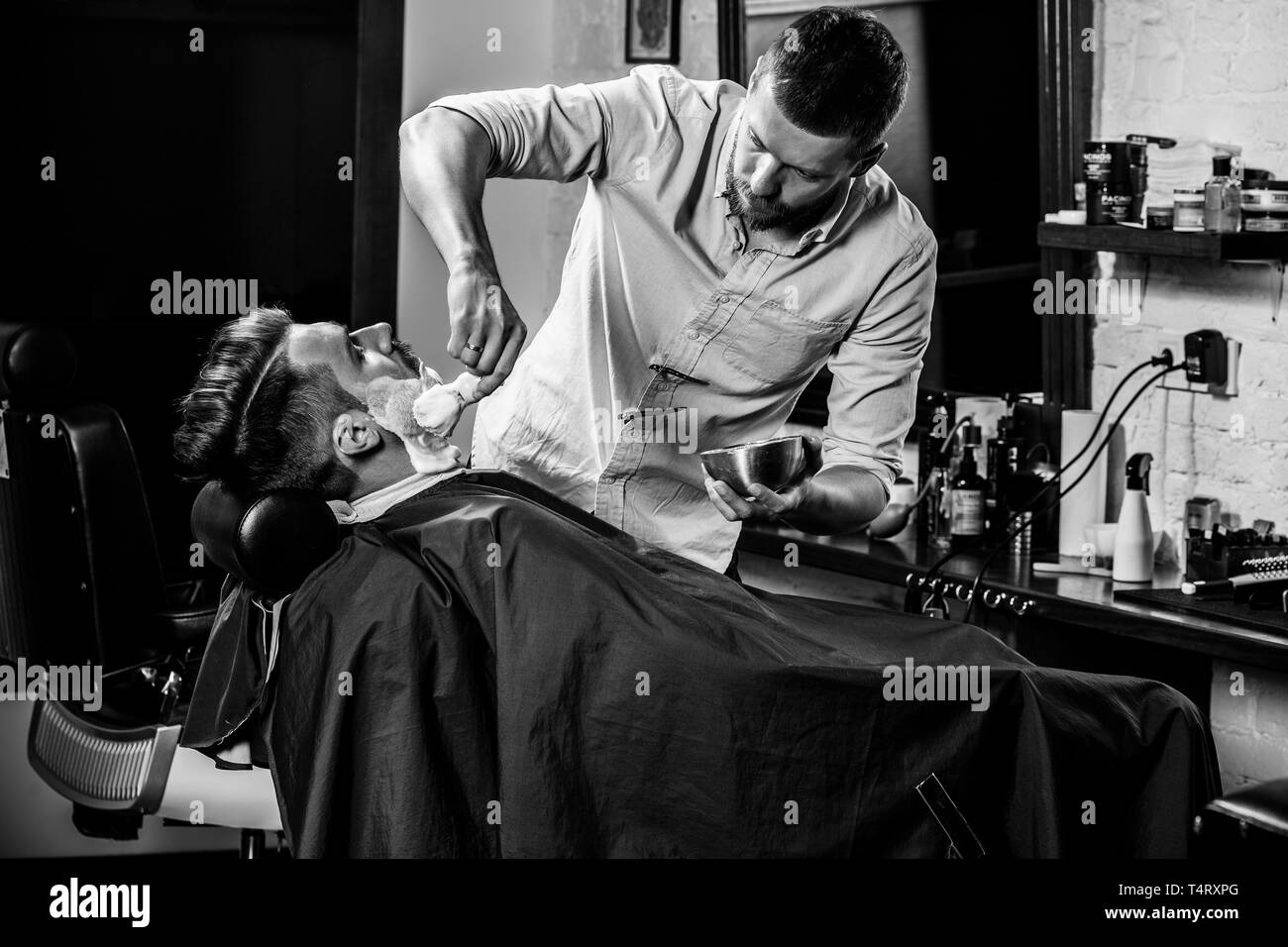 Client durante la rasatura della barba in barberia. Giovani bel barbiere e attraente uomo barbuto. Nero e bianco o incolore foto. Acconciatura, salone di bellezza e parrucchiere, barbiere, il concetto di stile di vita. Caucasian modelli maschili. Foto Stock