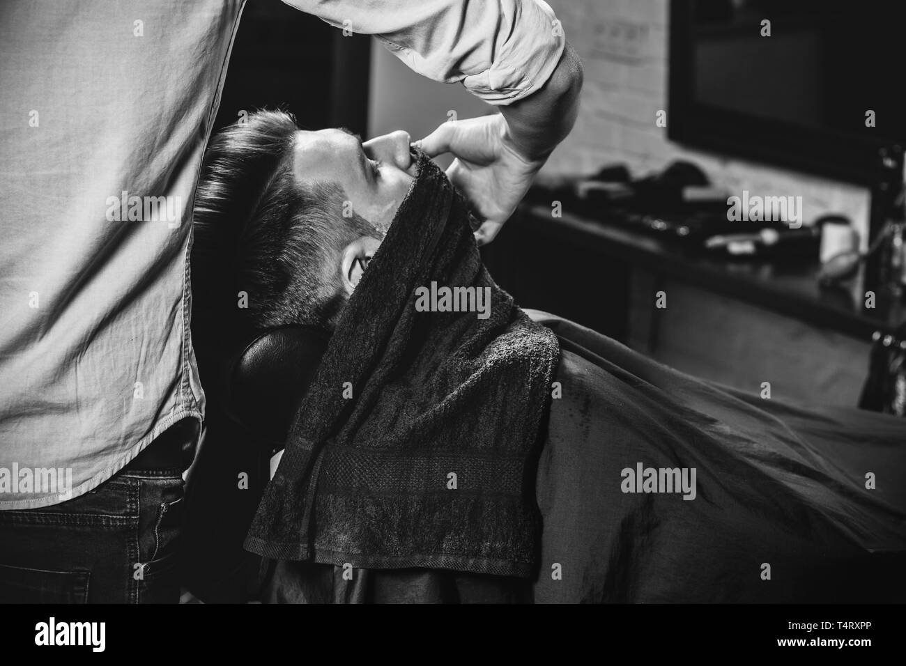 Client durante la rasatura della barba in barberia. Giovani bel barbiere e attraente uomo barbuto. Nero e bianco o incolore foto. Acconciatura, salone di bellezza e parrucchiere, barbiere, il concetto di stile di vita. Caucasian modelli maschili. Foto Stock