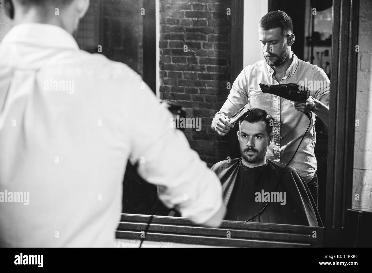 Giovani bel barbiere rendendo haircut per attraente uomo barbuto al barbiere. Nero e bianco o incolore foto. Acconciatura, salone di bellezza e parrucchiere, barbiere, il concetto di stile di vita. Caucasian modelli maschili. Foto Stock