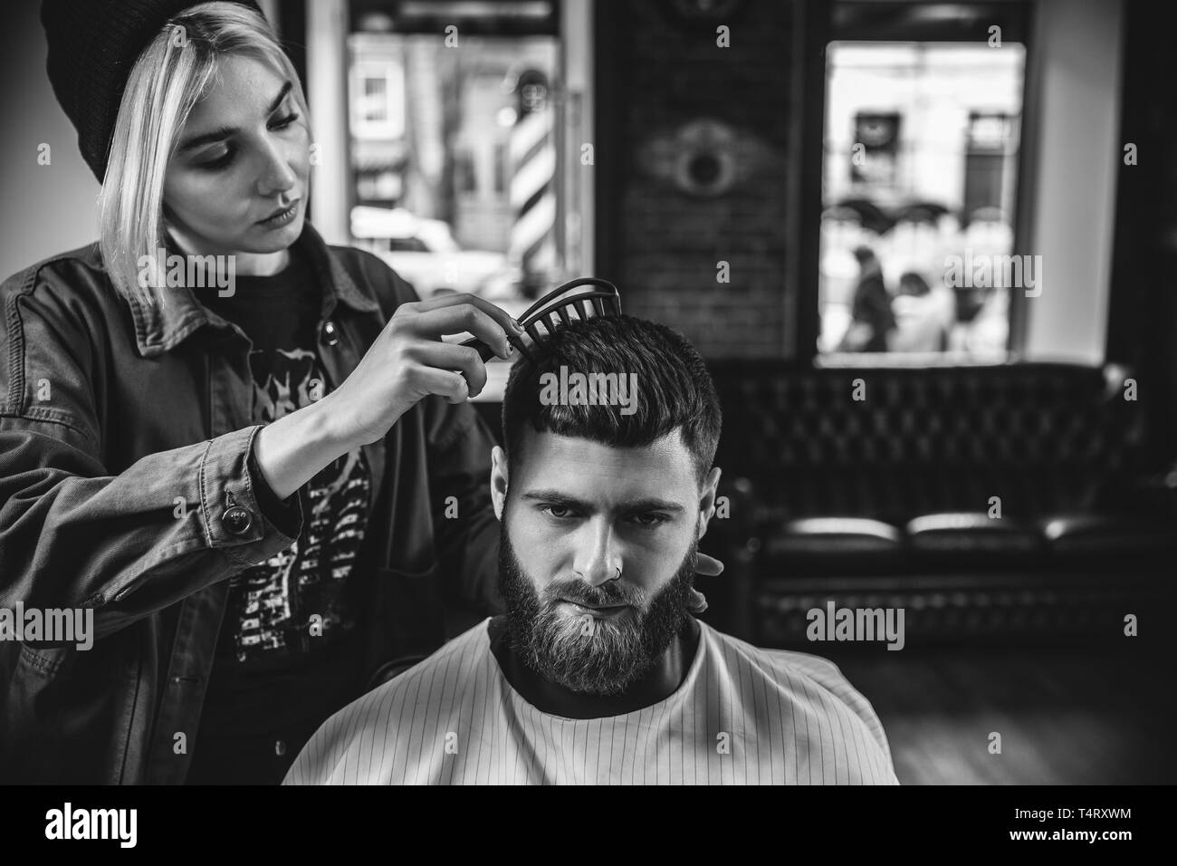 Giovane donna che fa il taglio di capelli uomo barbuto al barbiere. Barbiere femmina al salone. La parità tra i sessi. Donna nel maschio professione. Nero e bianco o incolore foto. Acconciatura, salone di bellezza e parrucchiere, il concetto di stile di vita. Foto Stock