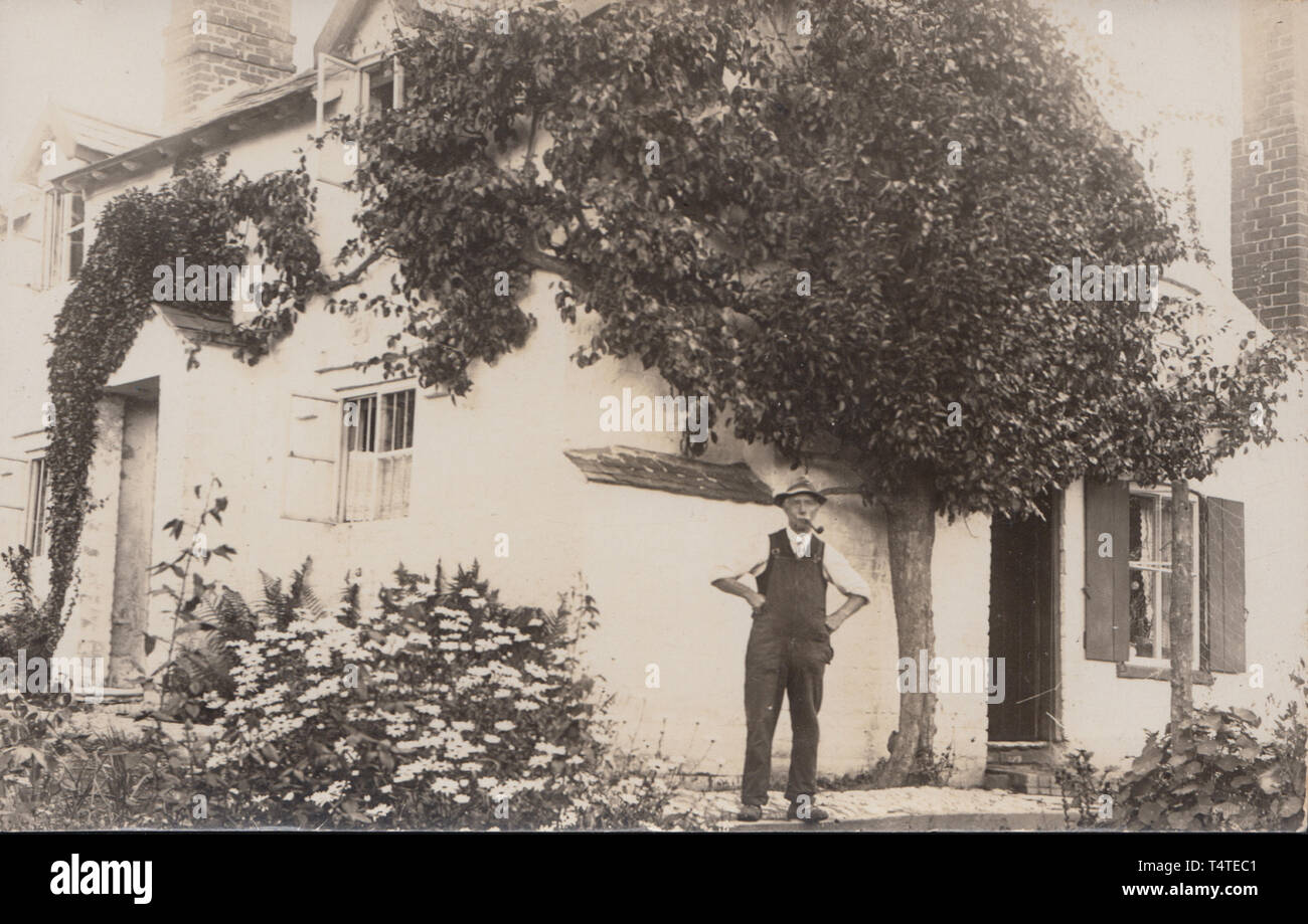 Vintage Cartolina fotografica che mostra una storica casa britannica. Uomo anziano si fermò al di fuori di fumare la pipa. Foto Stock