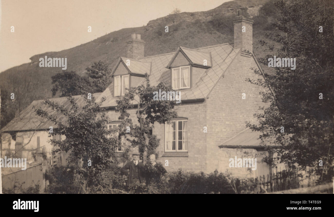 Vintage Cartolina fotografica che mostra una staccata storico britannico di casa in una posizione rurale ai piedi di una collina. Giovane si è levato in piedi nel giardino di proprietà. Foto Stock