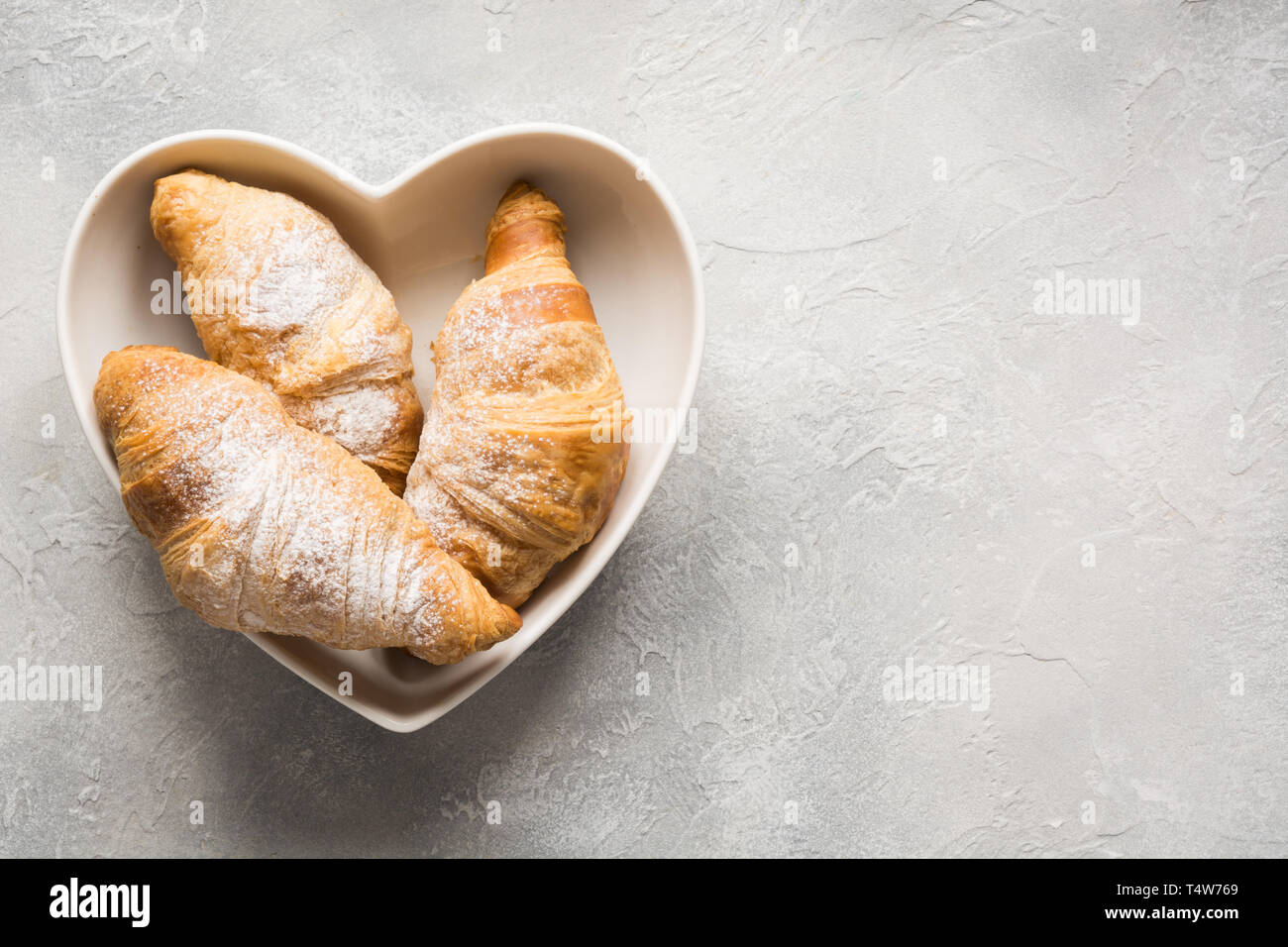 Croissant appena sfornati in piastra come cuore su sfondo grigio. Vista dall'alto. Copia dello spazio. Foto Stock