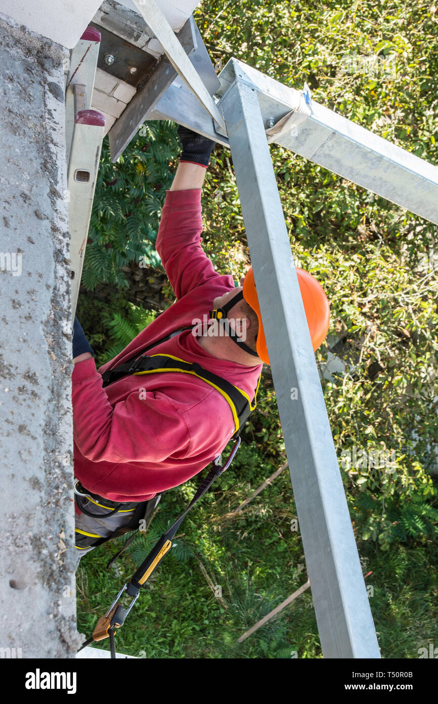 Altezza di lavoro. Intelaiatura metallica costruzione. Lavoratore manuale nel sistema di cavi del sedile. Workman con orange casco. Gruppo di balcone. Belaying sulla fune. Il dispositivo di protezione anticaduta. Foto Stock