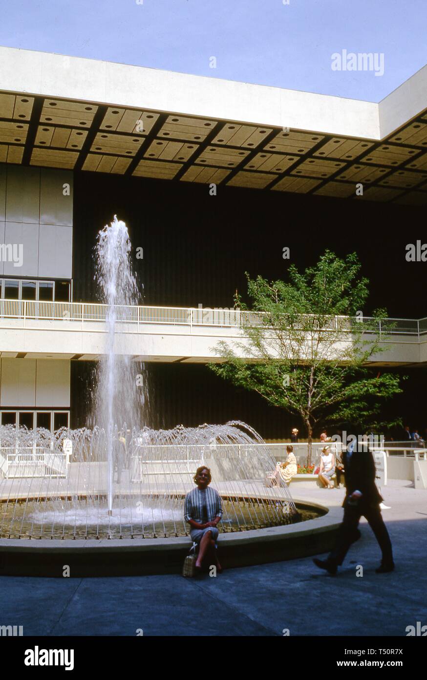 Un pedone maschio entra nel telaio, come una donna sorridente, seduto sul bordo di una fontana rotonda, pone di fronte a un padiglione espositivo, alla fiera mondiale di New York, Flushing Meadows Park, Queens, a New York, giugno 1964. () Foto Stock