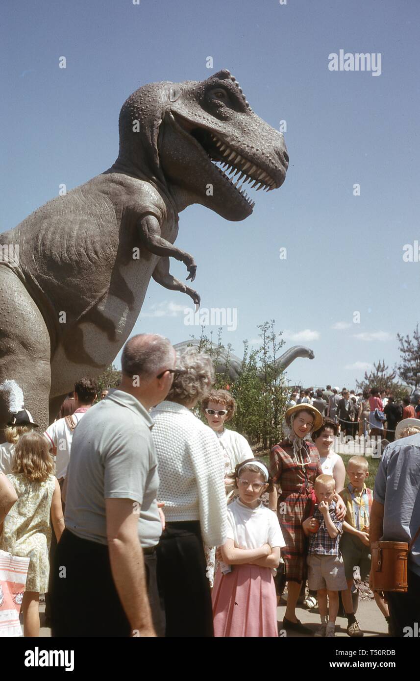 Una folla di gente che in una giornata di sole, a piedi e a porre in prossimità di un tessuto vetroresina Tyrannosaurus Rex al Dinoland presentano, Sinclair Pavilion, alla fiera mondiale di New York, Flushing Meadows Park, Queens, a New York, maggio, 1964. () Foto Stock