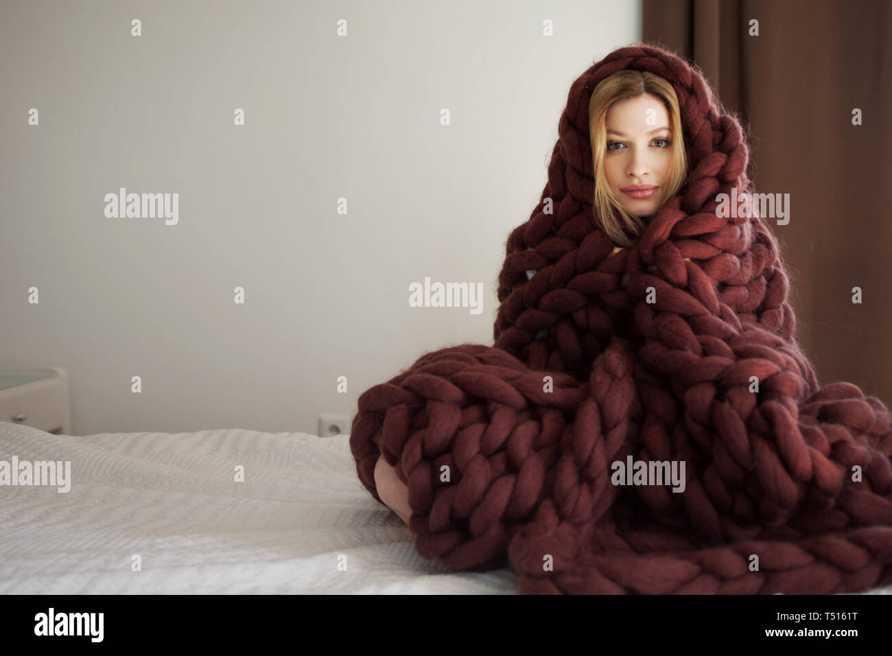 Carino giovane biondo a casa in camera da letto, seduta sul letto avvolto in una grande coperta di marrone. Una giovane donna di casa crogiolarsi in un accogliente coperta. Foto Stock