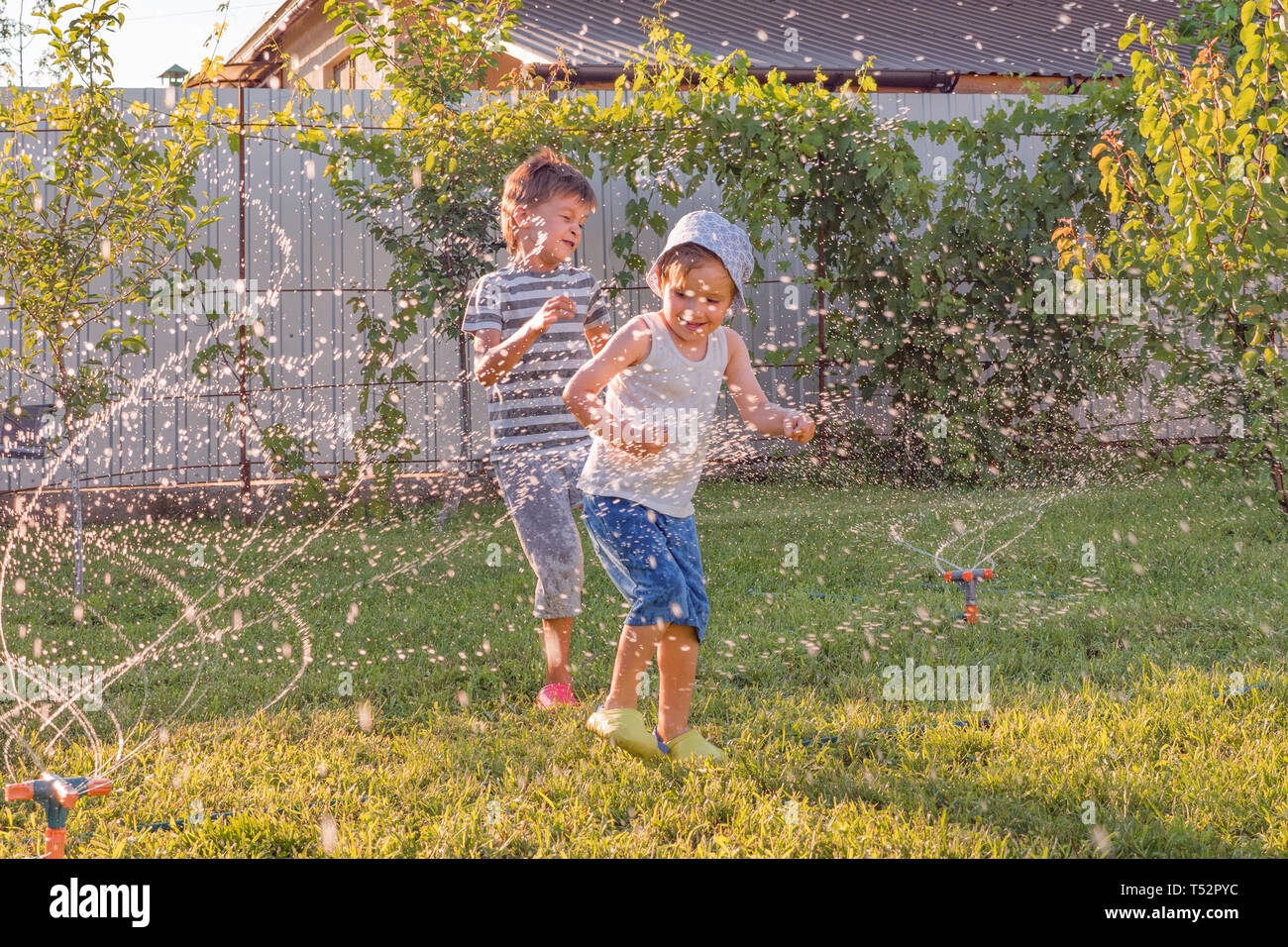 Attività estive. I bambini a giocare all'aperto con impianto automatico sistema di irrigazione. Ragazzo sorridente avendo divertimento all'aperto. I tubi flessibili di acqua di abbeveraggio l'erba Foto Stock