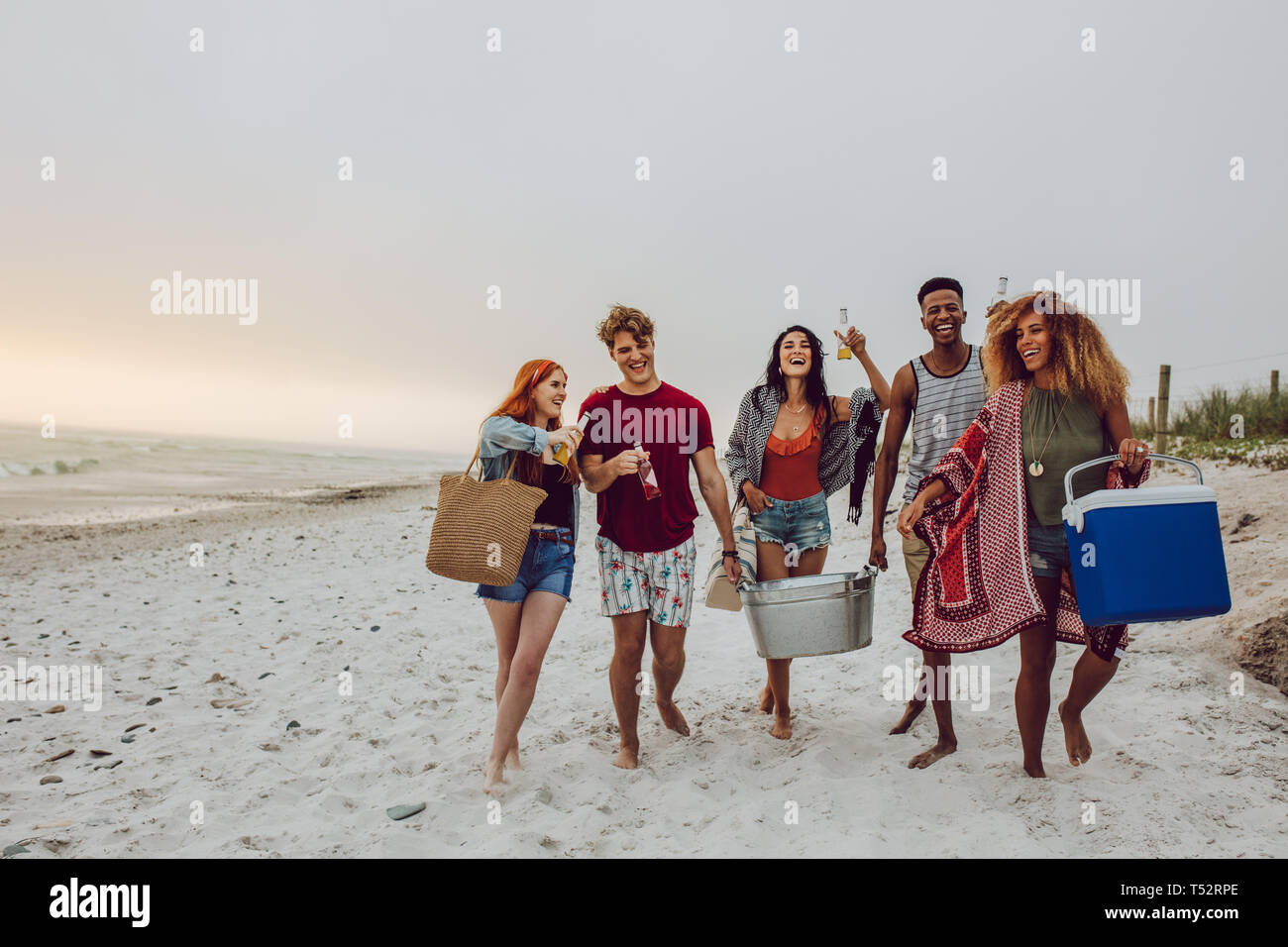 Un gruppo di giovani di camminare sulla spiaggia che porta una scatola del radiatore e vasca di bevanda. Giovani uomini e una donna sulla riva del mare. Foto Stock