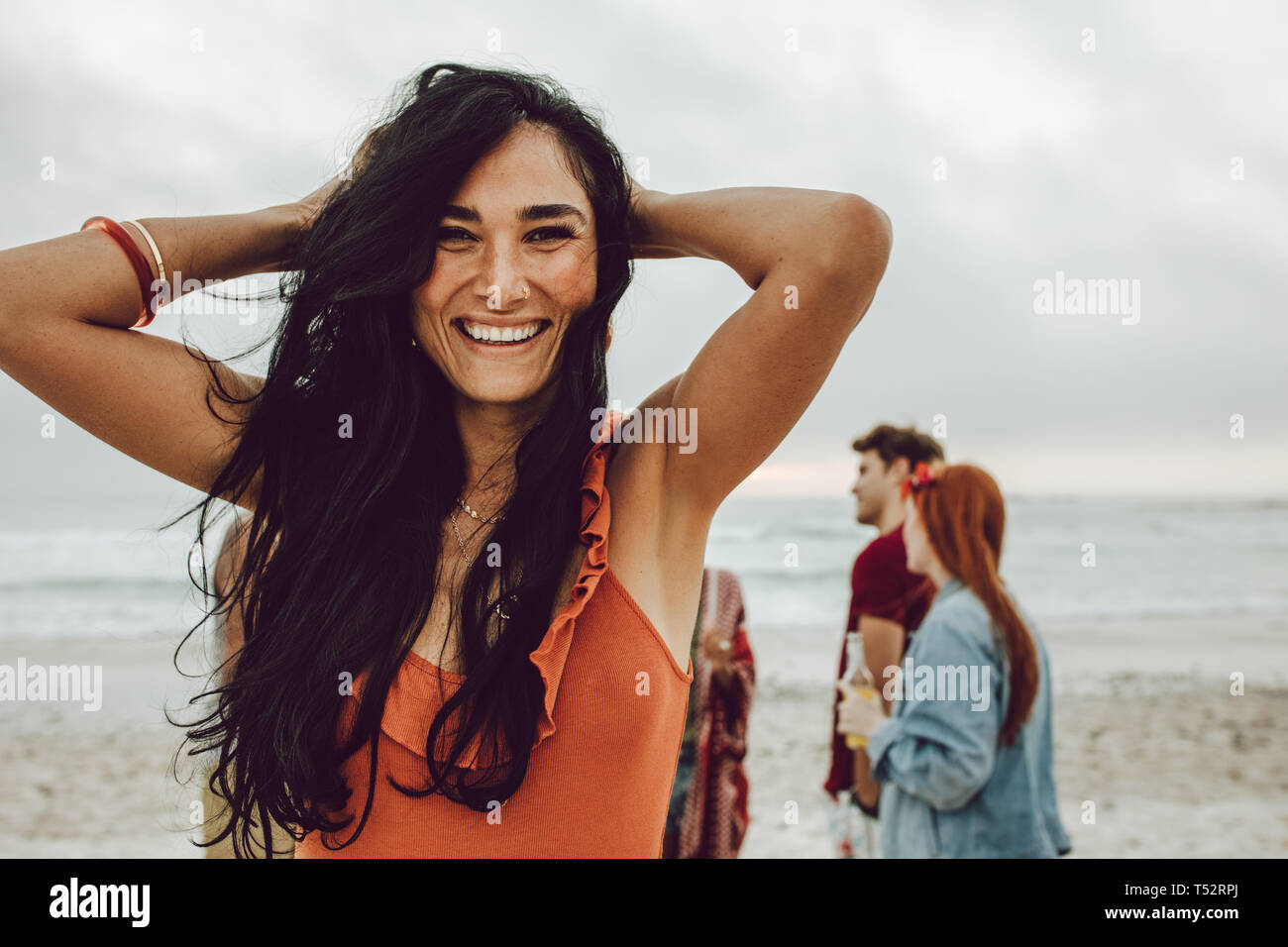 Bella giovane donna sulla spiaggia con gli amici all'indietro. Allegro femmina in casuals in piedi presso la spiaggia con un gruppo di amici in background. Foto Stock