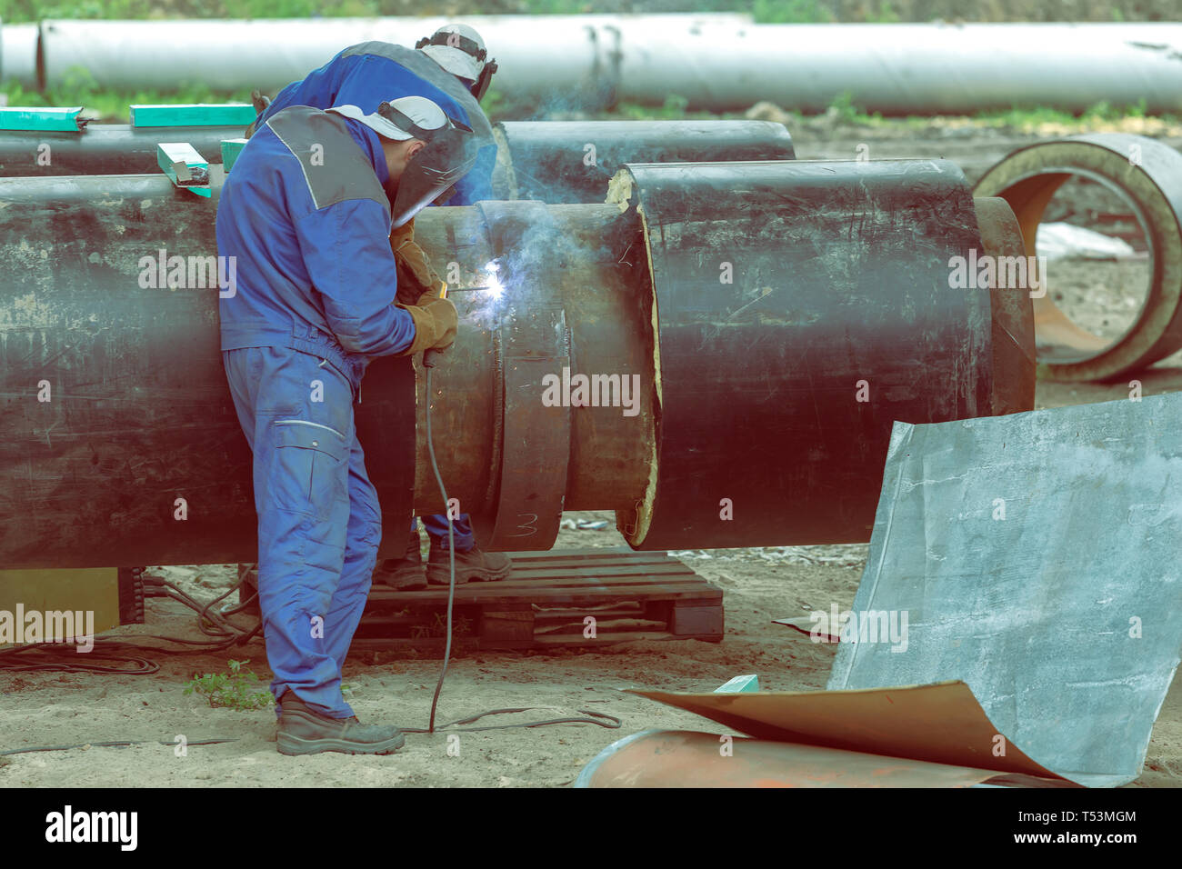Saldatrici giunti di saldatura su grandi isolati del distretto di acciaio tubo di riscaldamento con saldatura elettrica. Indossando gli ingranaggi di sicurezza. Effetto colore. Foto Stock