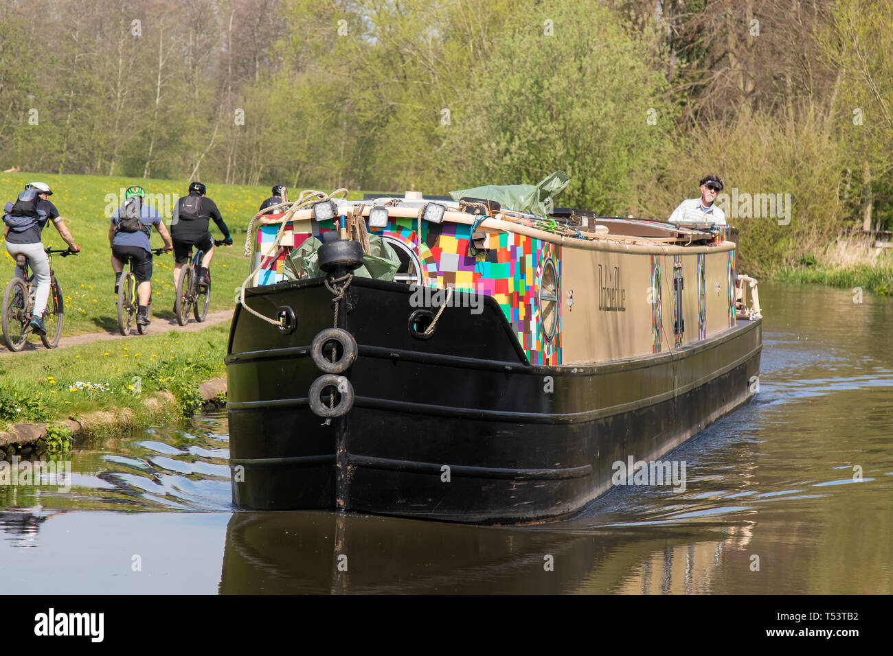 Primo piano del narrowboat in arrivo che viaggia sul canale britannico sotto il sole primaverile, con un solo operatore che gira sul retro. I ciclisti percorrono il canale. Foto Stock