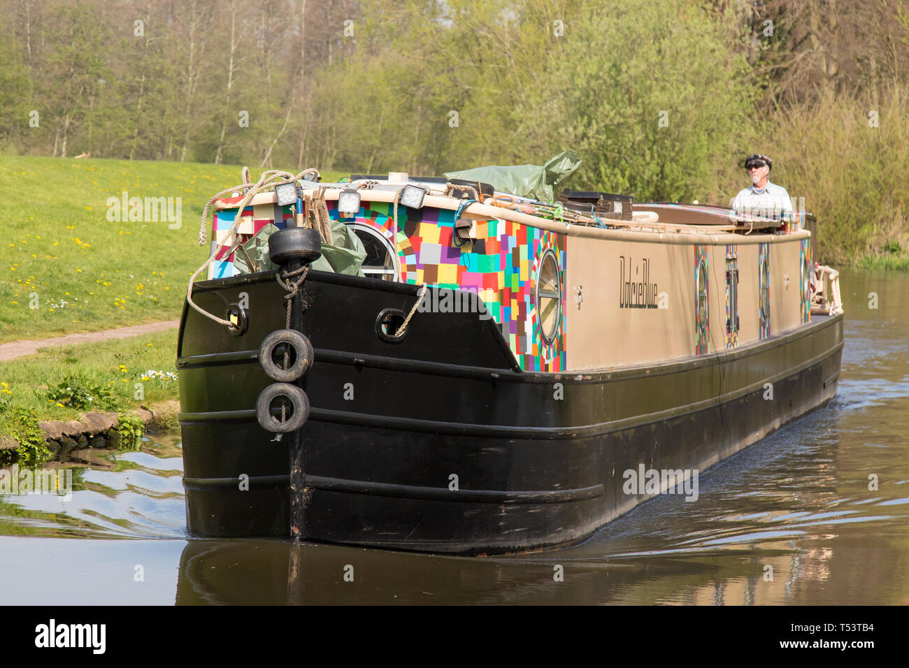 Paesaggio primo piano di una barca stretta in arrivo che viaggia sul canale britannico sotto il sole primaverile; uomo isolato e in pensione in cappellino si trova sul canale posteriore della barca. Foto Stock