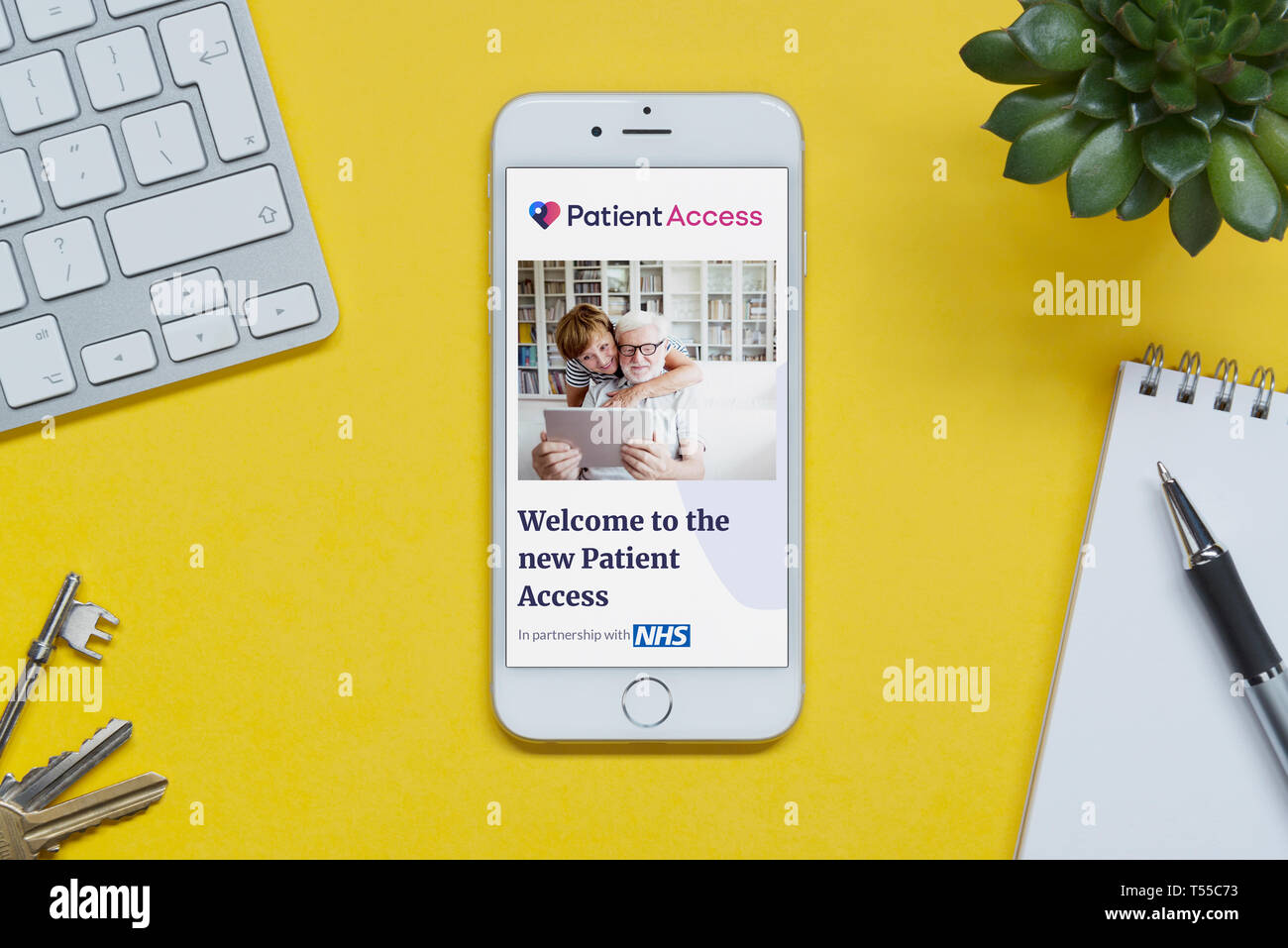 Un iPhone che mostra il paziente sito web Accesso poggia su uno sfondo giallo tabella con una tastiera, tasti, blocco note e di impianto (solo uso editoriale). Foto Stock