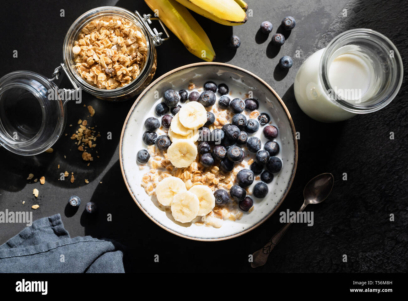 Sana colazione oat cereali con latte, mirtillo e banana nera su sfondo di calcestruzzo. Tabella vista superiore, luce cruda fotografia di cibo Foto Stock