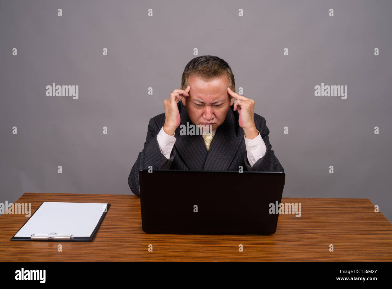 Maturo imprenditore asiatico seduto con un tavolo di legno contro il grigio Foto Stock