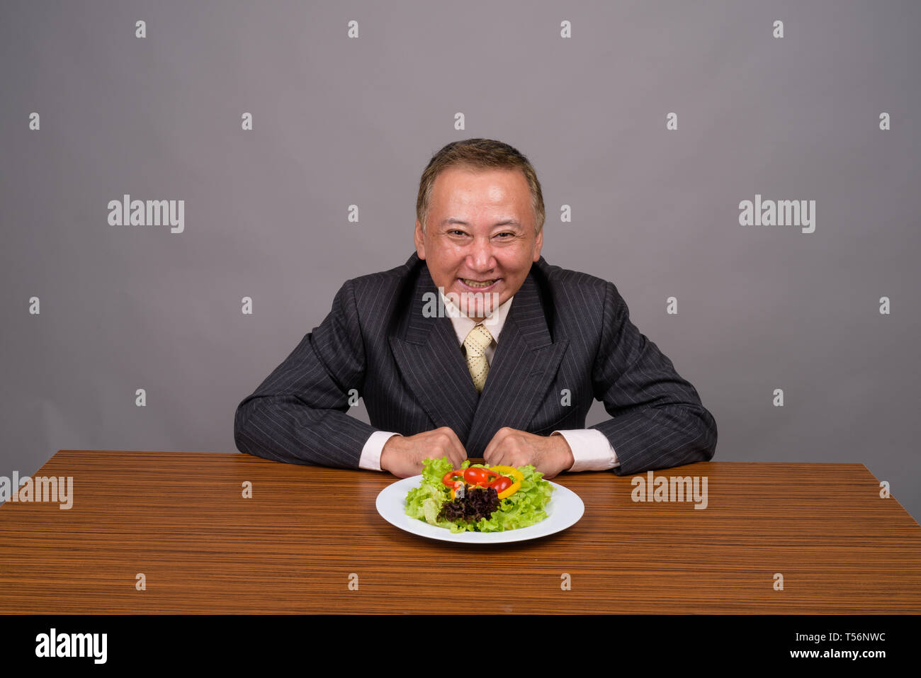 Maturo imprenditore asiatico seduto con un tavolo di legno contro il grigio Foto Stock