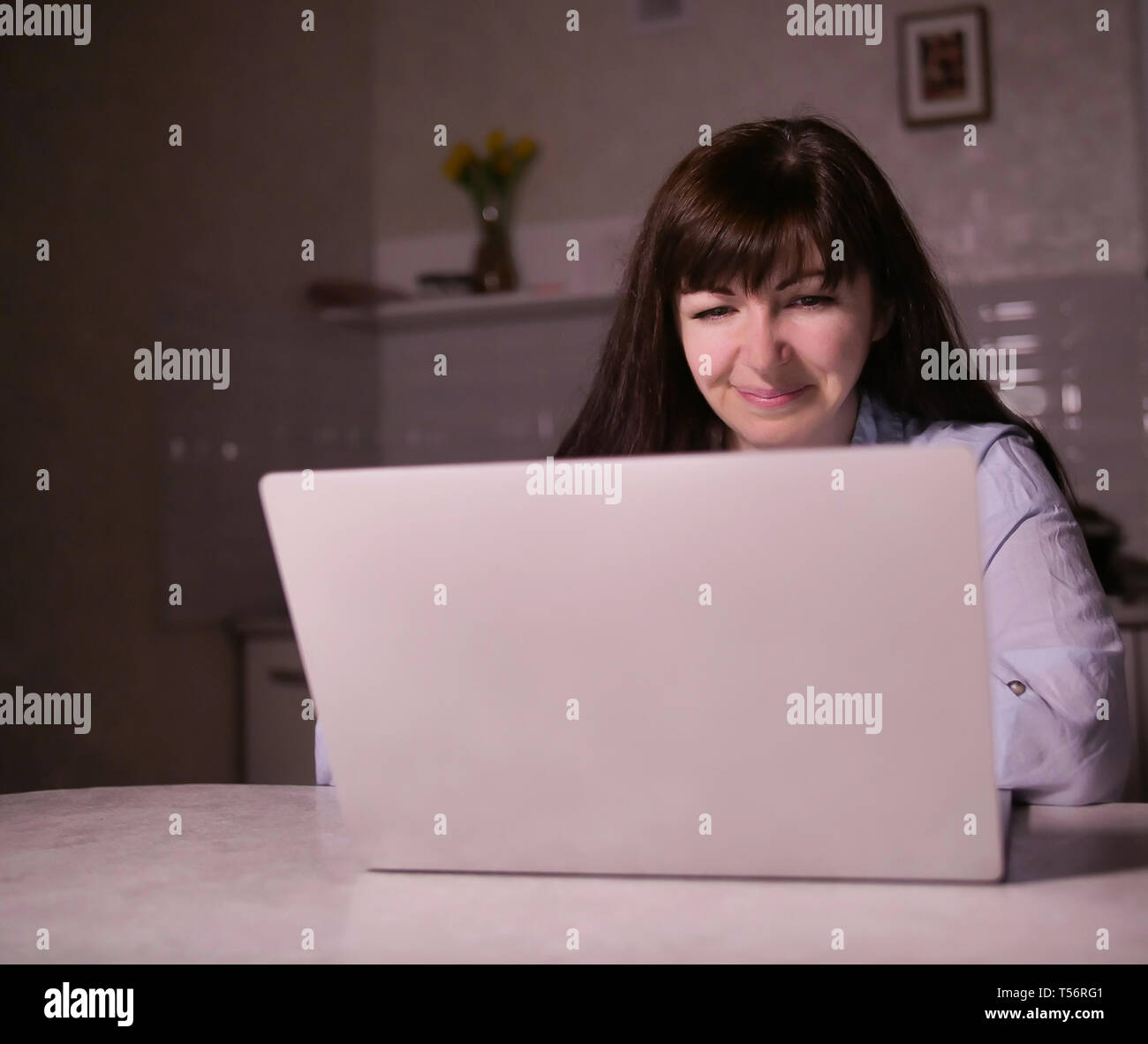 Sorridente bruna giovane donna seduta in serata nella sua cucina con un laptop Foto Stock