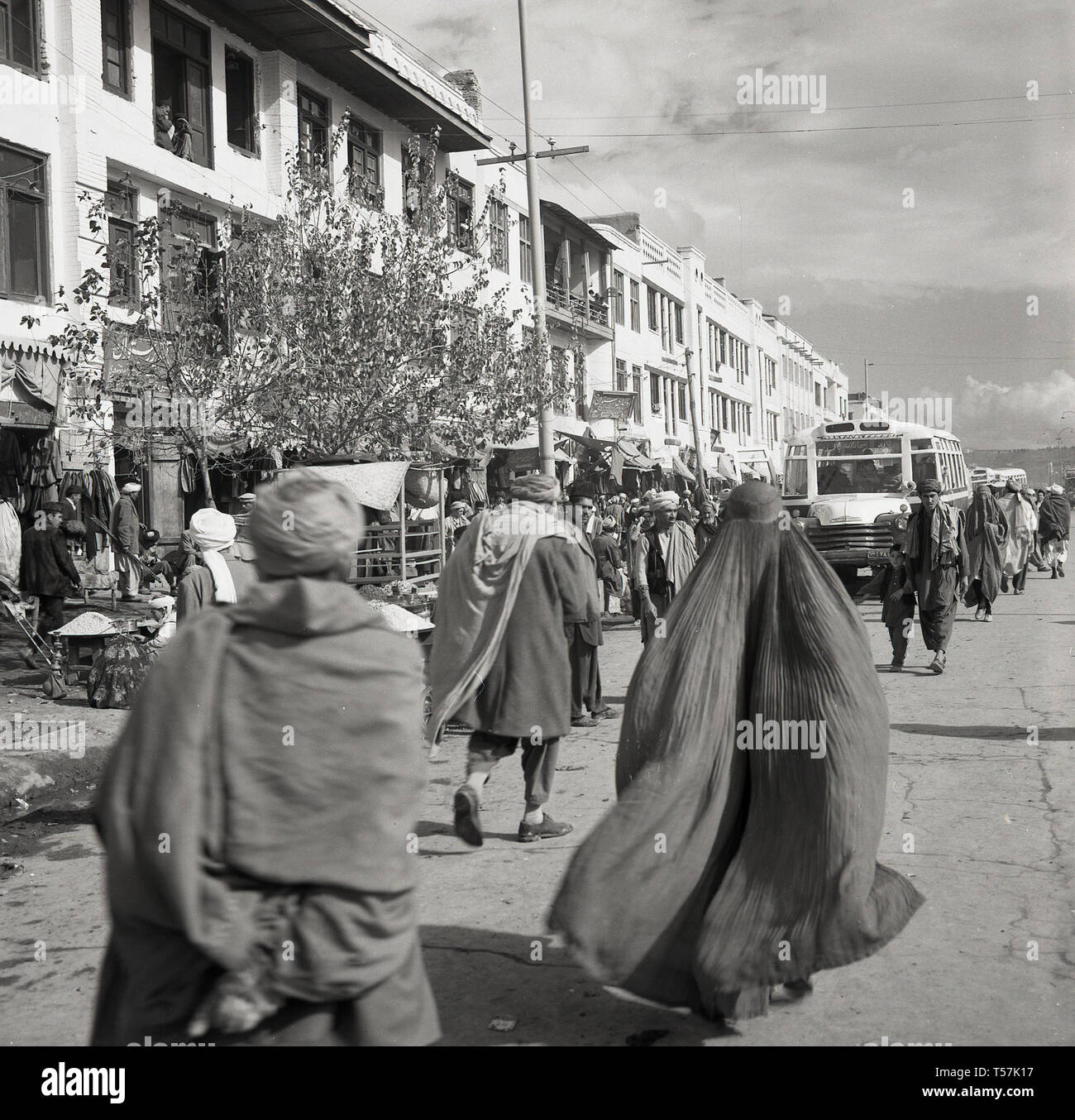 Degli anni Cinquanta, Kabul, Afghanistan, locali afghani in abito nazionale in una ampia strada polverosa passando le bancarelle del mercato e il bus locale nel centro della citta'. Foto Stock