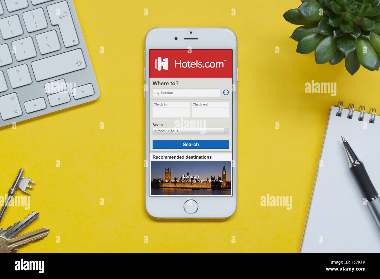 Un iPhone che mostra il sito web hotels.com poggia su uno sfondo giallo tabella con una tastiera, tasti, blocco note e di impianto (solo uso editoriale). Foto Stock