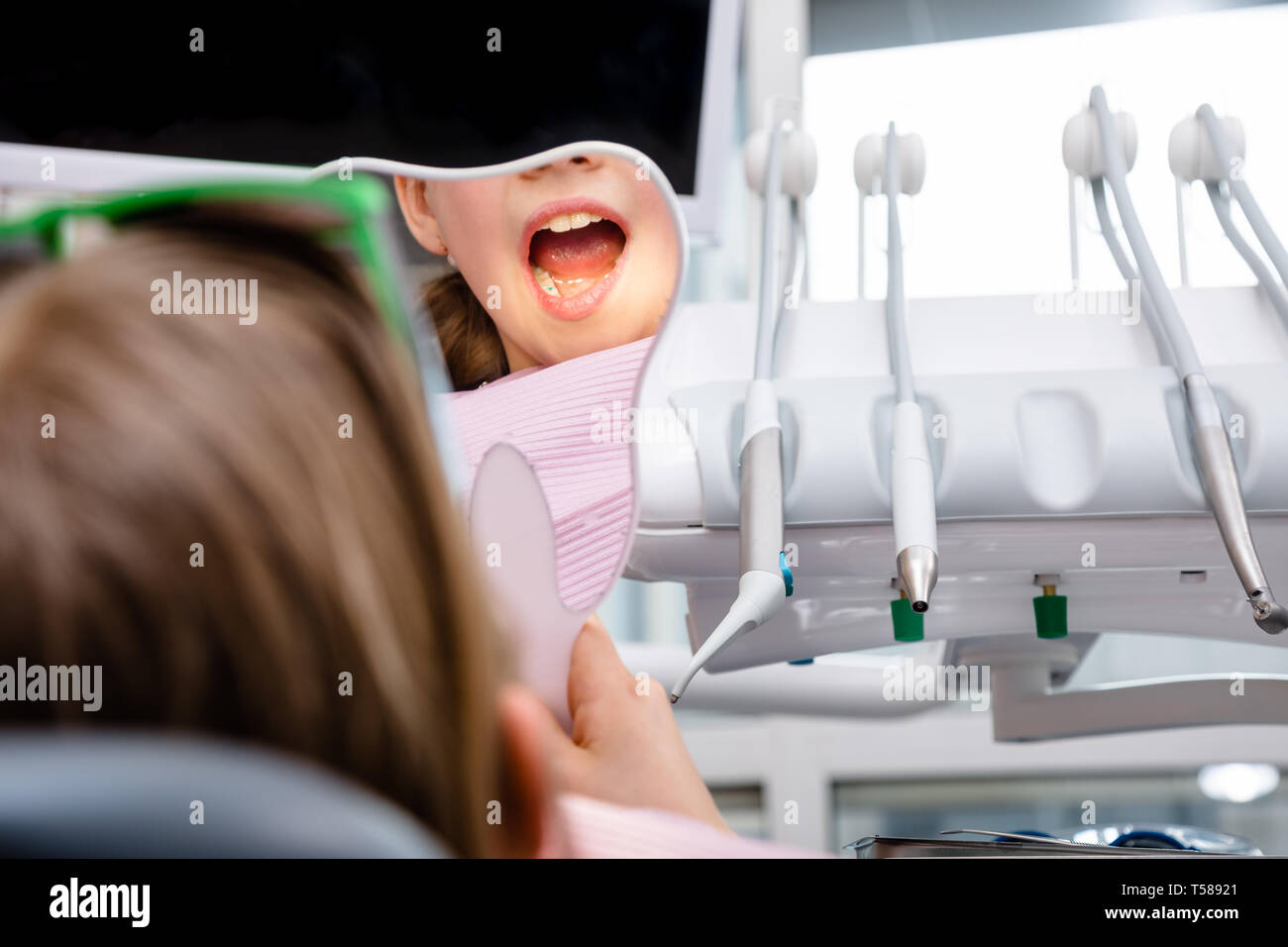 Preteen ragazza seduta in un riunito dentale di ispezionare i denti riparati guardando a forma di dente specchio in pazienti pediatrici clinica dentale Foto Stock