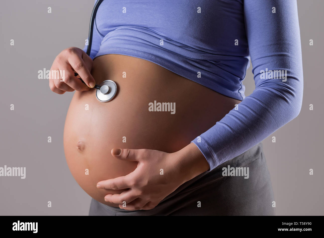 Immagine della donna incinta di ascolto heartbeat di bambino con uno stetoscopio su sfondo grigio. Foto Stock