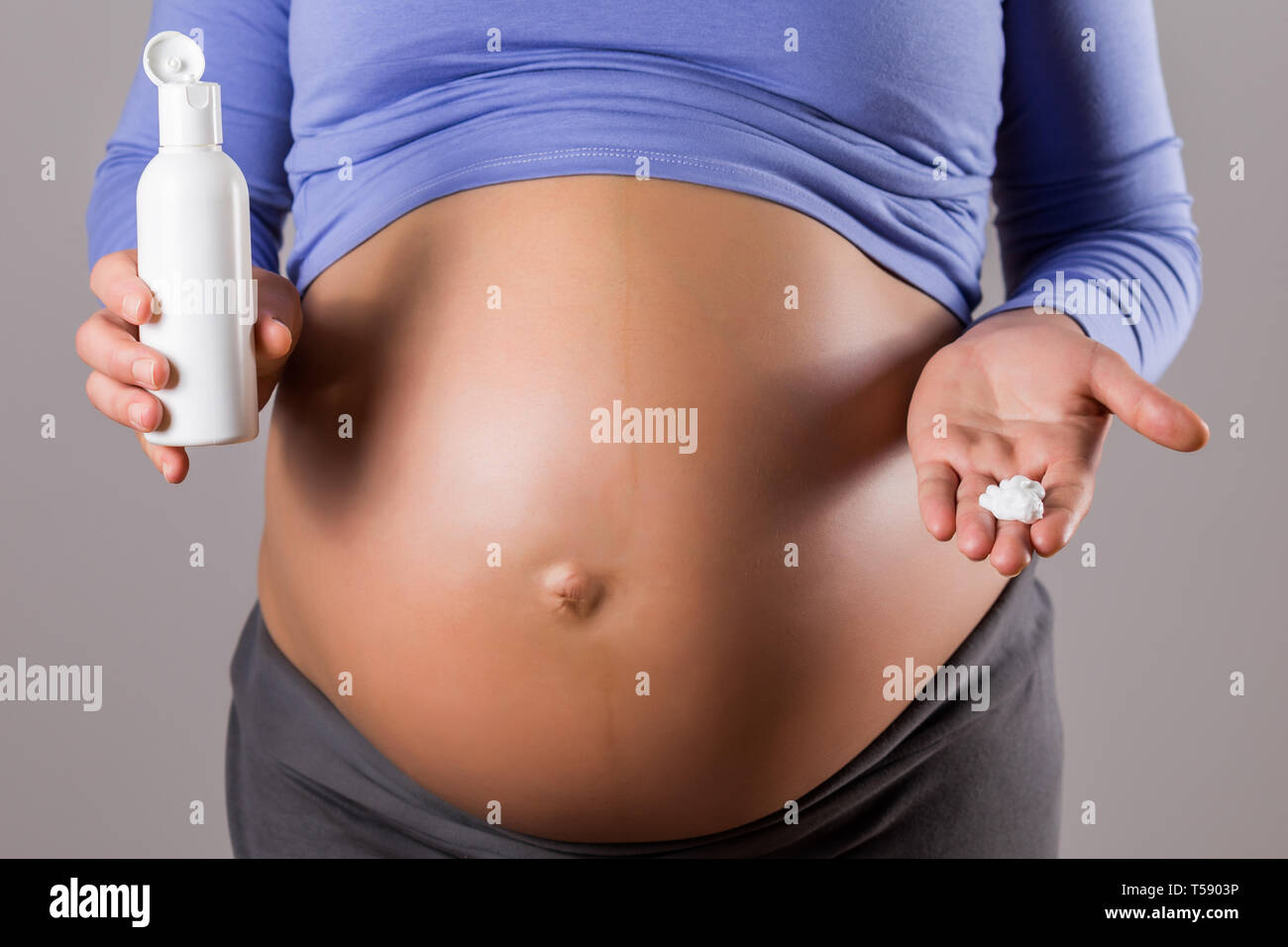 Immagine della donna incinta di applicare una crema idratante sul suo stomaco su sfondo grigio. Foto Stock