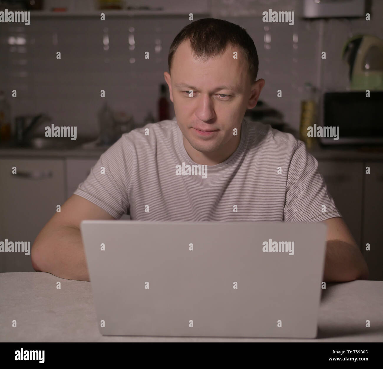 Attraente giovane uomo seduto in serata in cucina con un laptop Foto Stock
