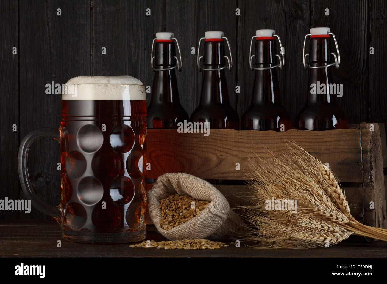 Bicchiere da birra sul tavolo con cassa piena di bottiglie, con la produzione di birra di orzo e frumento mazzetto, scuro dello sfondo in legno Foto Stock