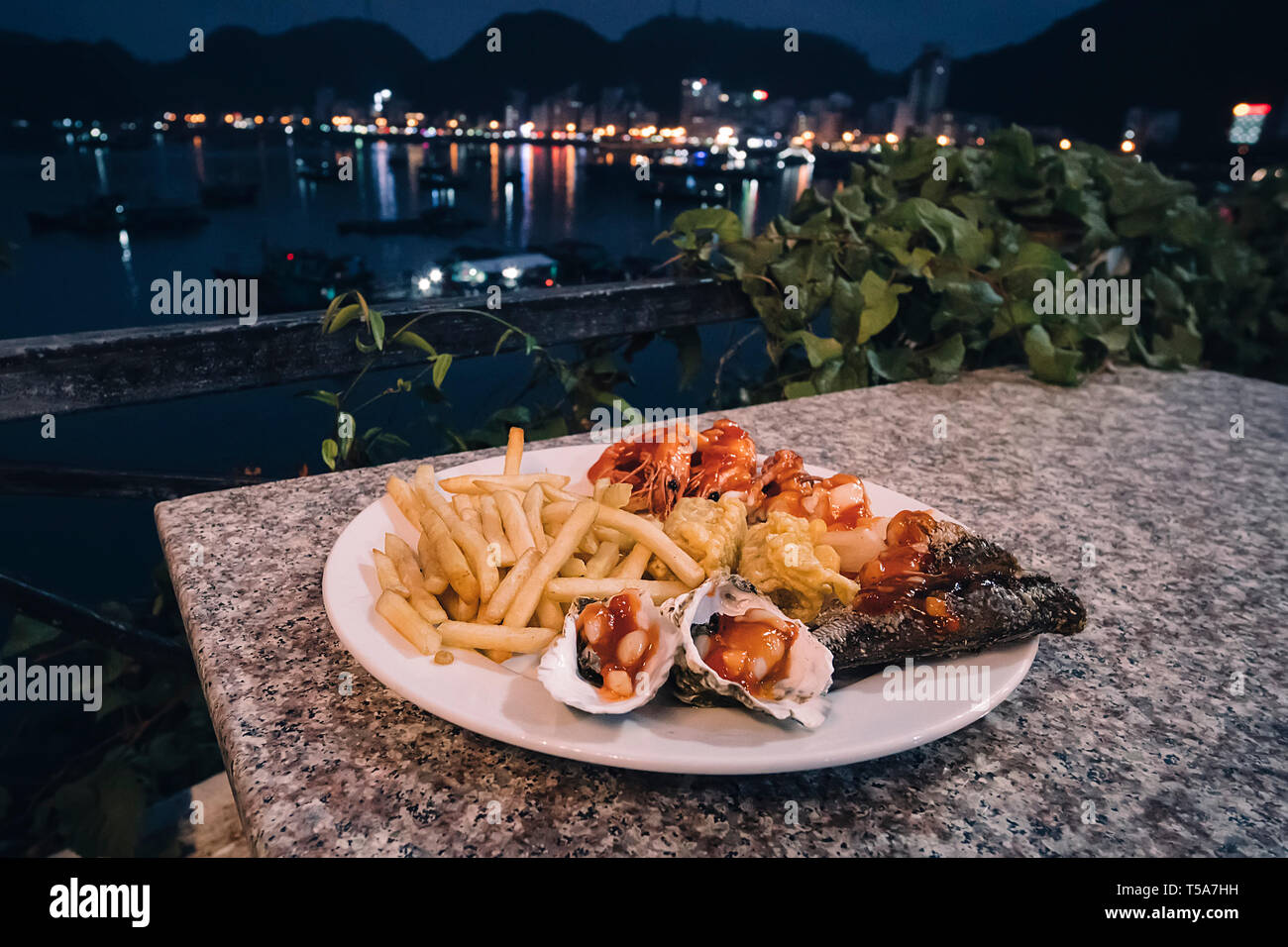 Piastra con frutti di mare, cozze, gamberi, pesce, polpi e patatine fritte sul tavolo contro il mare di notte all'aperto. Il Vietnam la Baia di Ha Long Foto Stock