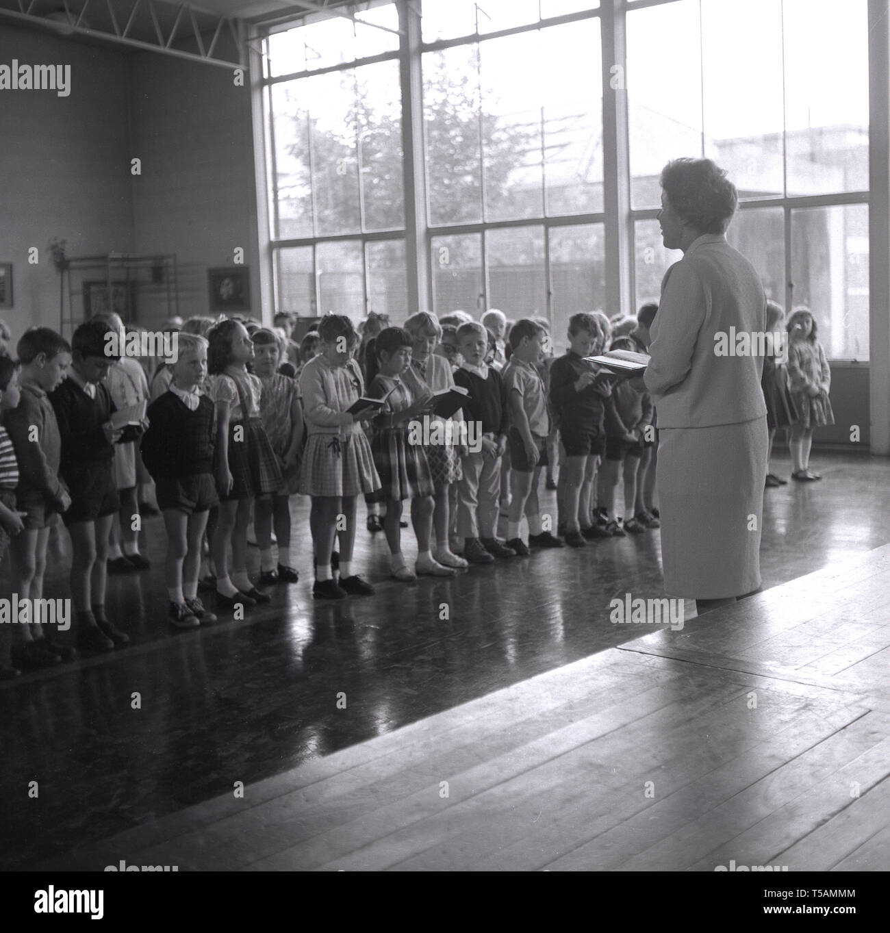 Anni sessanta, storica direttrice la lettura di un brano da un libro - eventualmente la bibbia - ad un gruppo di bambini della scuola primaria in piedi in una linea di assemblaggio di mattina, Inghilterra, Regno Unito. Foto Stock