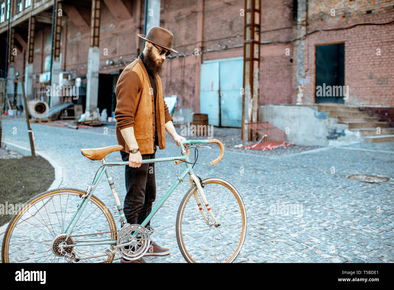 Stile di vita ritratto di un barbuto hipster vestito elegantemente camminando con retro bicicletta sul industriale di background urbano Foto Stock
