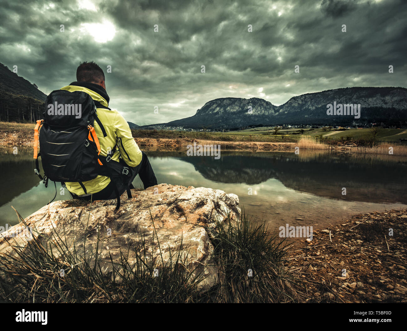 Una persona si siede sul bordo di una scogliera che si affaccia su un azzurro lago alpino in un giorno nuvoloso Foto Stock