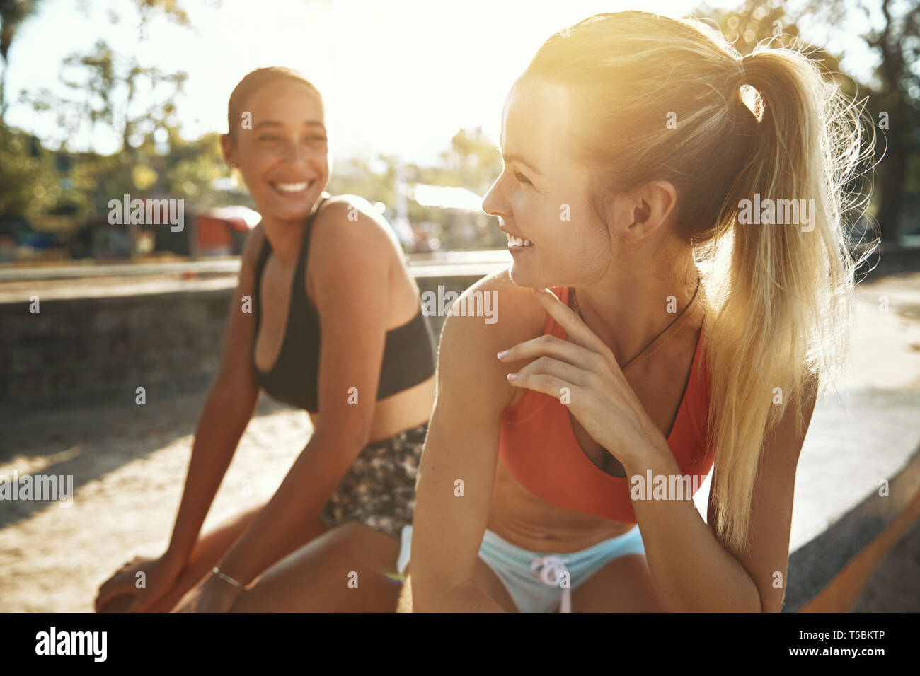 Montare due giovani donne in sportswear sorridere mentre seduti insieme al di fuori in una giornata di sole prendendo una pausa dal loro allenamento Foto Stock