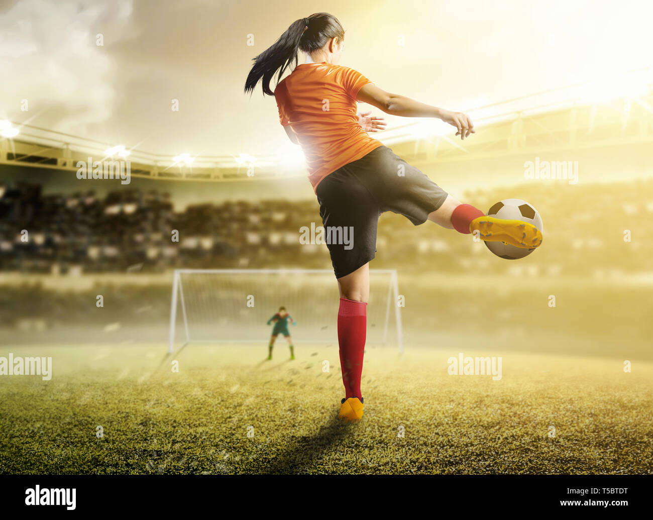 Vista posteriore di asian football player donna in maglia arancione calciare il pallone in pena casella sul campo di calcio a Stadium Foto Stock