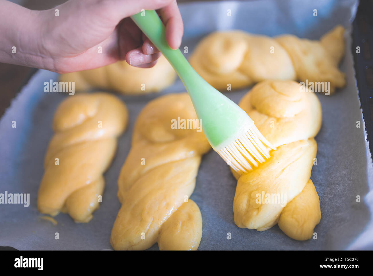 Close-up di mano intrecciato di vetratura dolci panini lievito utilizzando silicone spazzola di pasta prima della cottura Foto Stock