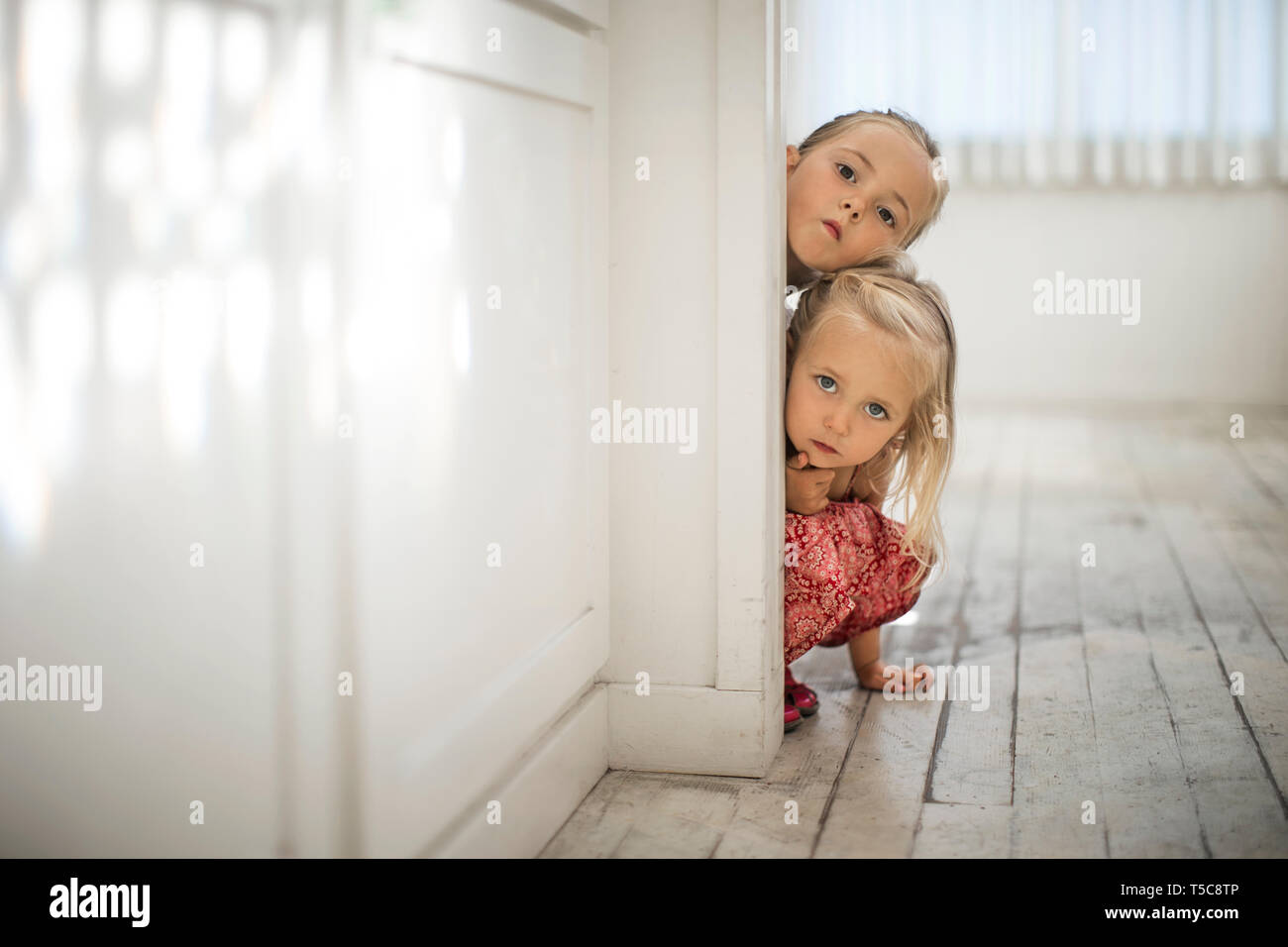 Due giovani sorelle bionda peek intorno al bordo di un infisso di una porta. Foto Stock