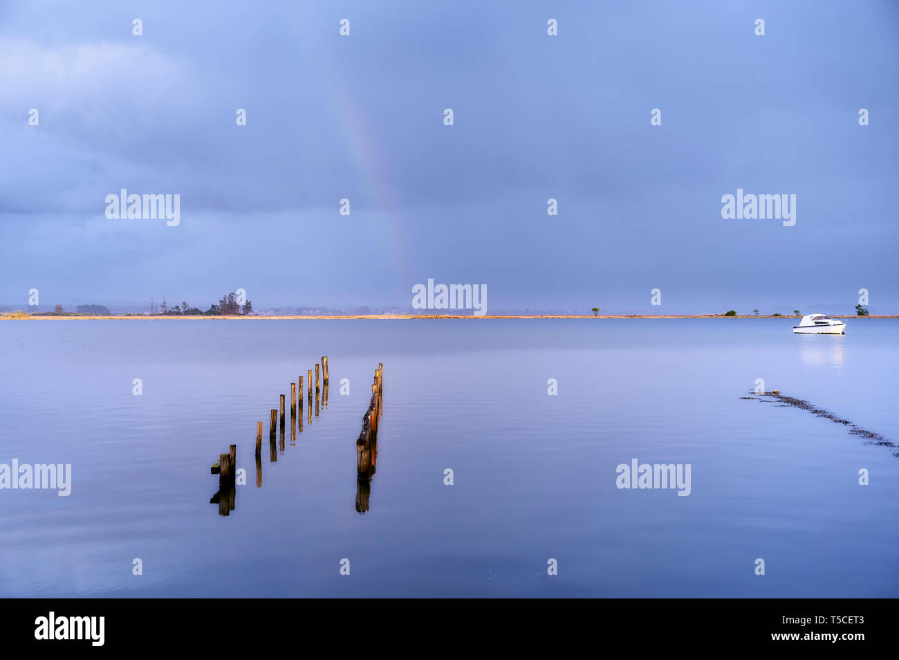 Picchetti di antiche quays, illuminata dal sole, segnare un percorso sulle acque blu attraverso il lago fino al Rainbow sulla sponda opposta con nuvole scure. Foto Stock