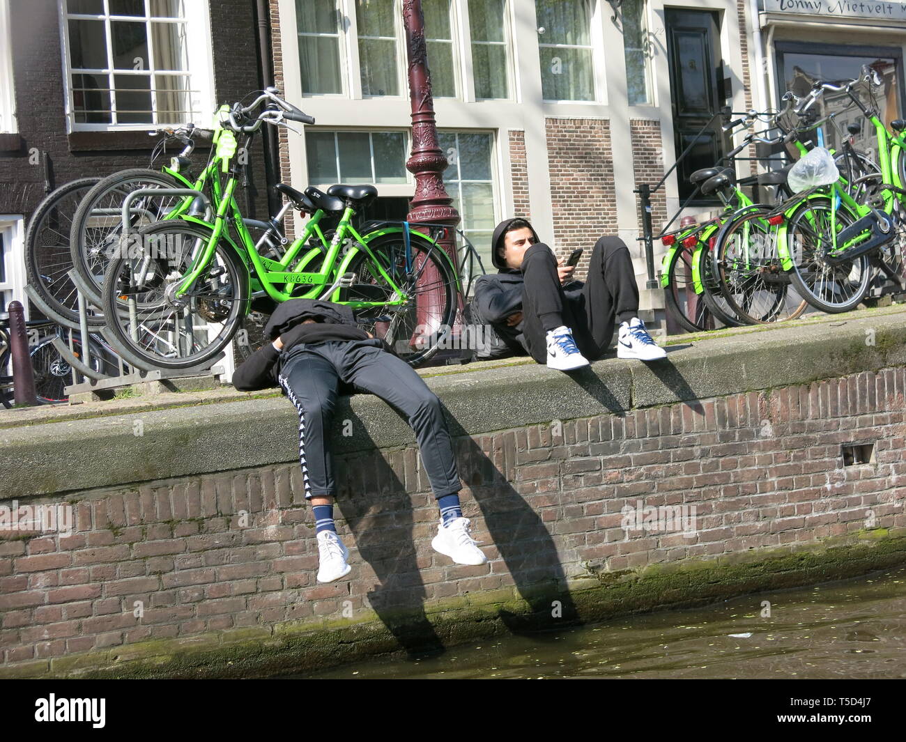 Due giovani in abito da adolescente, in hoodies, formatori e jeans, giacciono sulle rive del canale di Amsterdam in un assolato pomeriggio di aprile Foto Stock