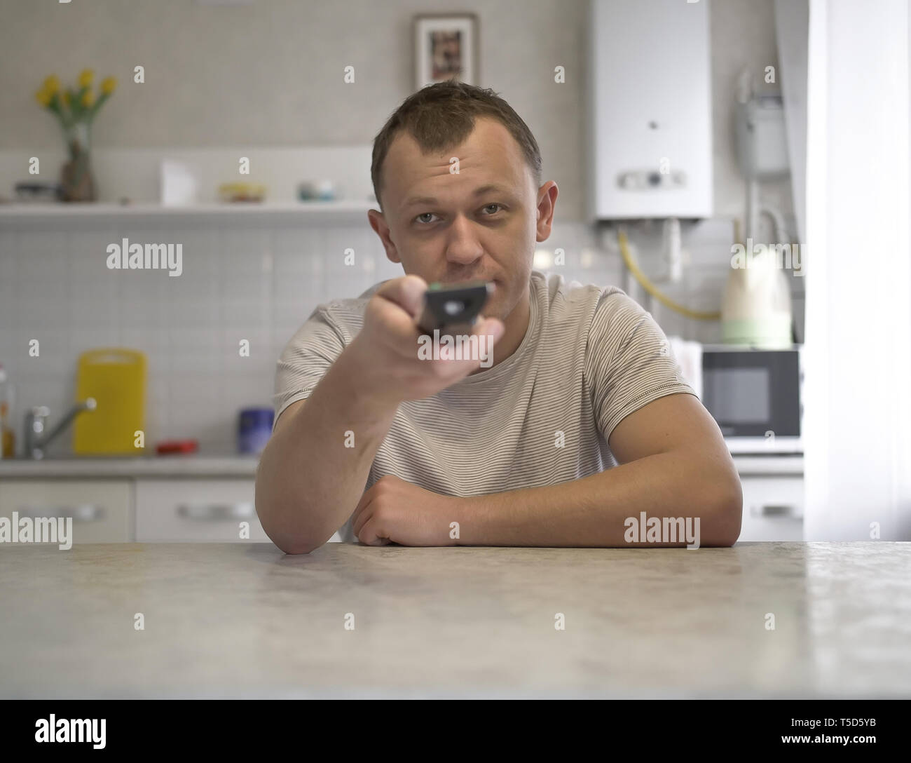 Giovane ragazzo con un telecomando TV interruttori di controllo di canali durante la seduta in cucina, vista frontale Foto Stock