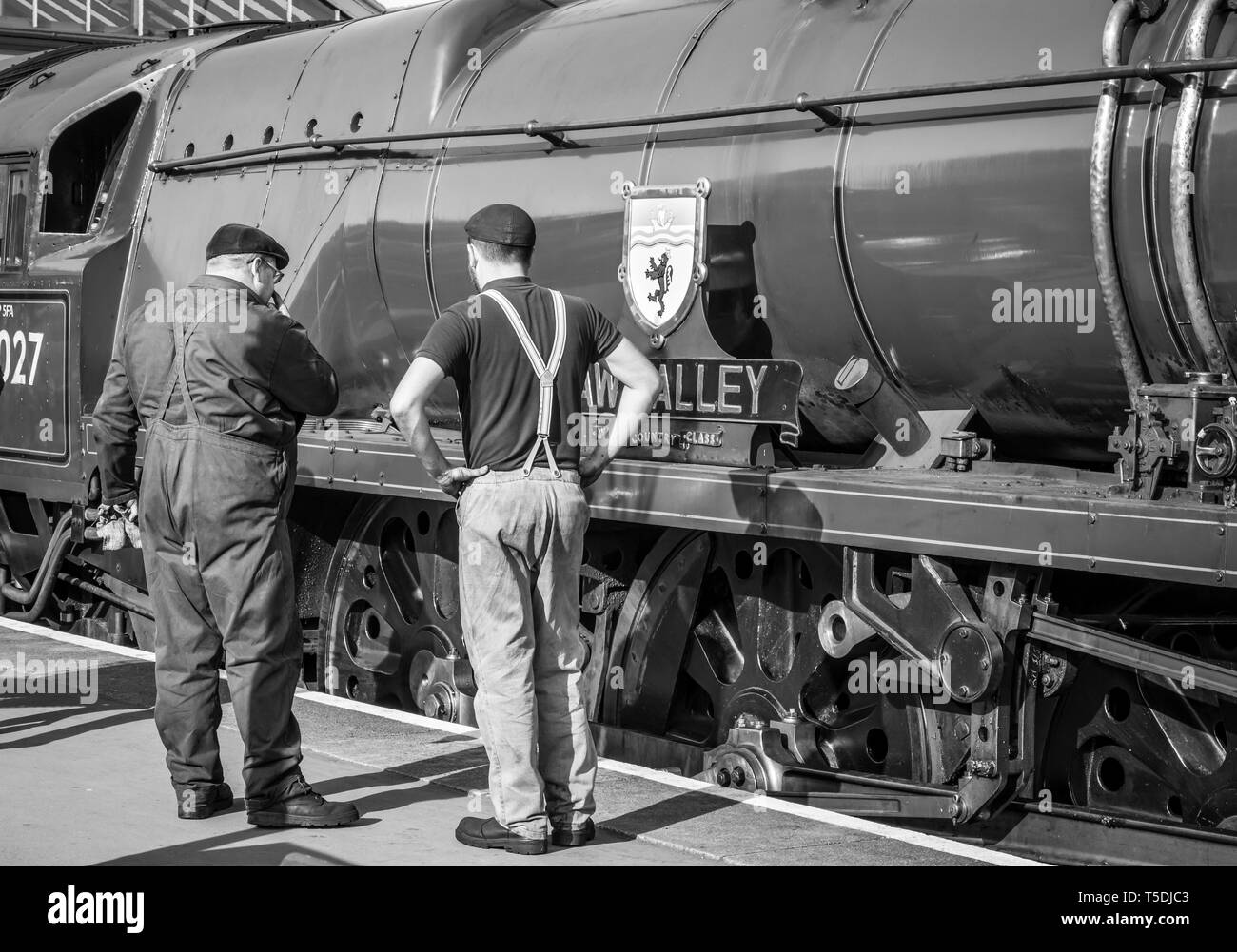 Vista posteriore in bianco e nero dell'equipaggio della locomotiva a vapore in piedi sulla piattaforma ferroviaria al sole che ispeziona il lato fisso del motore vintage del Regno Unito. Foto Stock