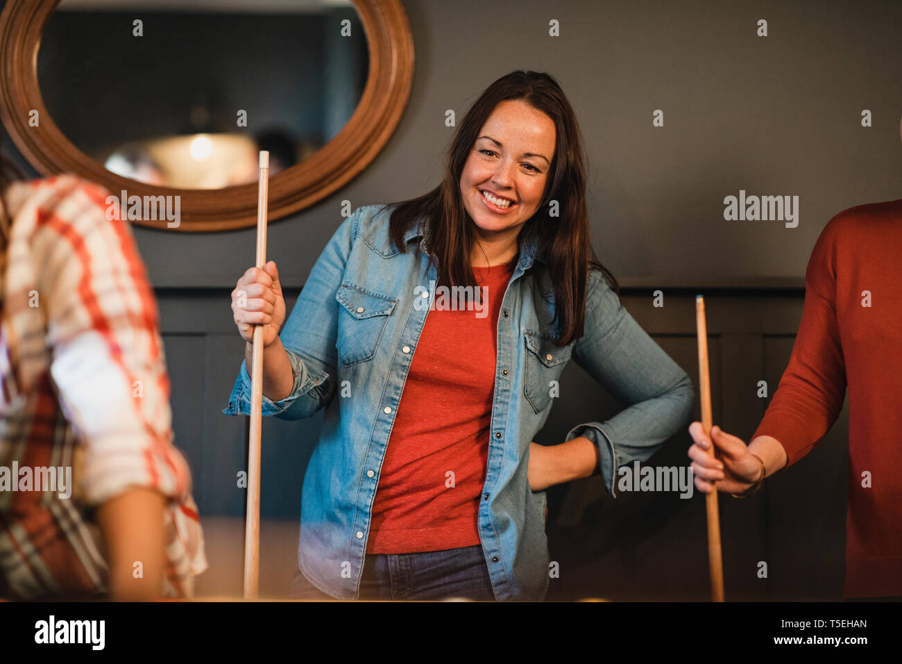 Metà donna adulta in piedi in ambienti chiusi durante la riproduzione di un gioco di piscina. Ella è in possesso di un pool cue e guardare il gioco in attesa del suo andare. Foto Stock