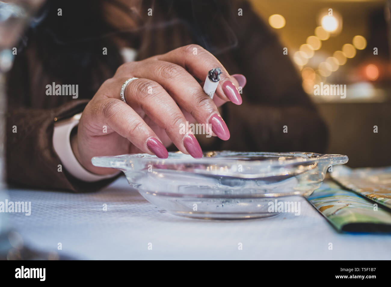 Origine asiatica donna con unghie verniciate, tenendo in mano una sigaretta su un posacenere in un bar. Foto Stock