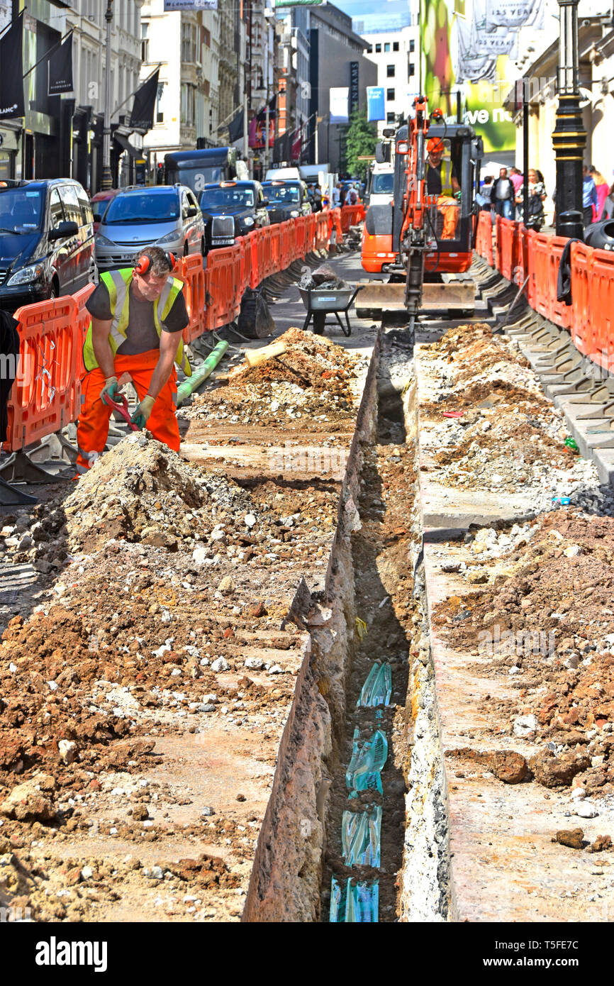 Inceppamento di traffico e lavori stradali contraente trincea di scavo in strade e vie London West End per cavi utilizzando mini escavatore dietro barriere Londra Inghilterra REGNO UNITO Foto Stock