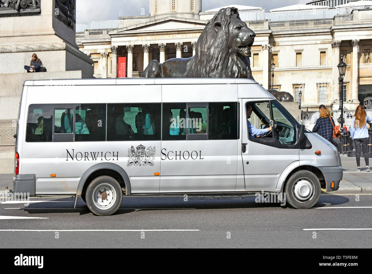 Scena di strada laterale del privato ed indipendente Scuola Norwich mini bus & driver sul viaggio a Londra guidando passato Lion statua in Trafalgar Square England Regno Unito Foto Stock
