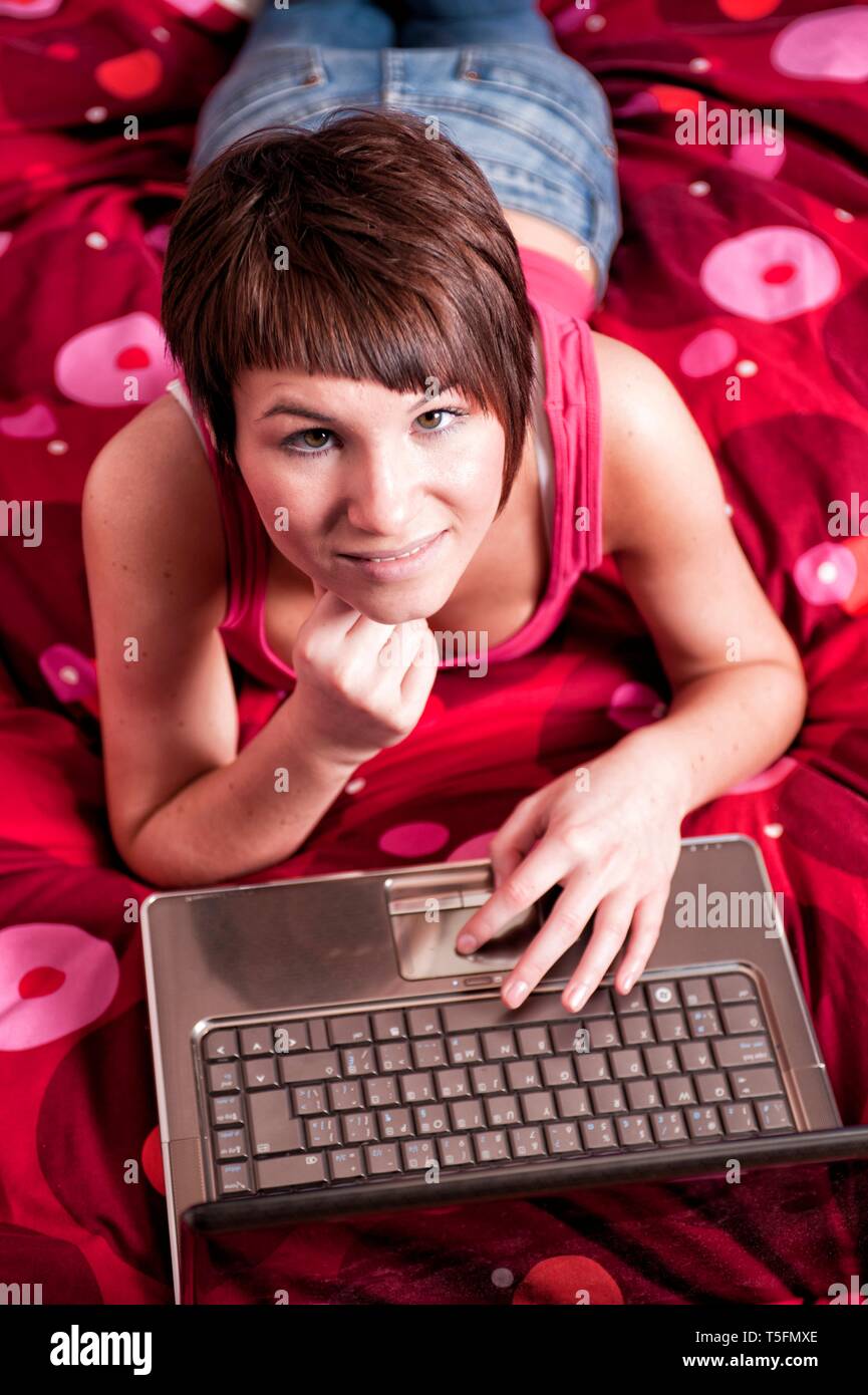 Junge Frau liegt auf ihrem Bett Mit einem Leptop vor sich vor sich und schaut in die freundlich in die Kamera. Foto Stock